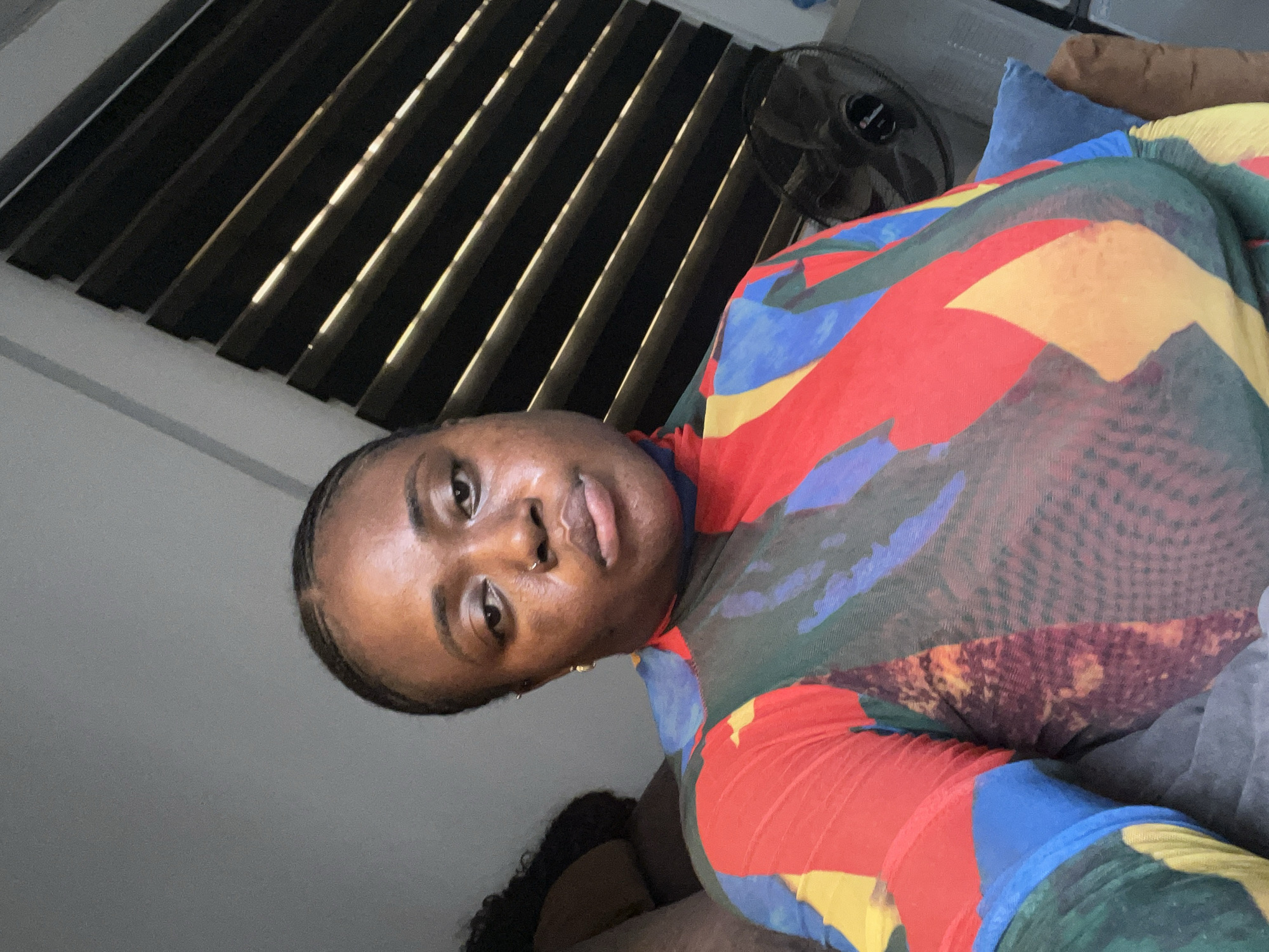 A medium shot of a person with dark skin and a nose ring, wearing a multicolored, geometric-patterned top. The person is sitting or lying down on a bed with blue and gray pillows. A black fan is visible in the background, along with window blinds. The location is potentially in Abuja, Nigeria, but it is difficult to definitively confirm without further information.