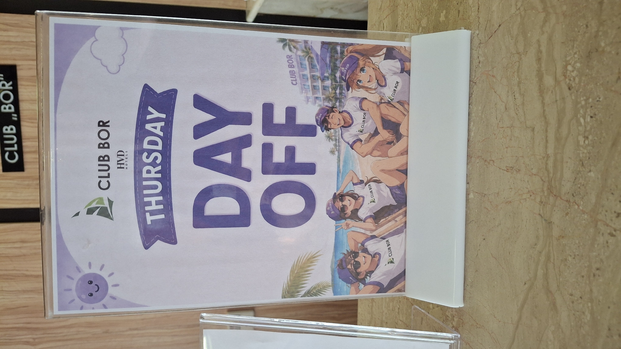 A framed sign for "CLUB BOR" is displayed on a marble countertop. The sign features a cartoon illustration of four young adults at a beach, with a resort in the background. Text on the sign reads "THURSDAY DAY OFF". The top of the sign has a sun icon and the "CLUB BOR" logo, along with "HVD HOTELS". The background appears to be a hotel lobby. The sign is supported by a white plastic stand.