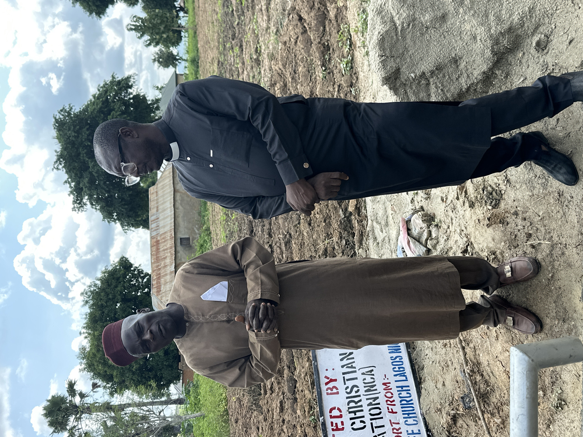 Two men are standing outdoors in Afogo, Nigeria. The man on the left is wearing a tan, knee-length robe with a white pocket square, a red kufi hat, and brown loafers. He is looking at the camera with his hands clasped in front of him. The man on the right is wearing a black cassock with a white clerical collar and black shoes. He is looking off to the side. In the background, there is a dirt field, some trees, and a building with a corrugated metal roof. A sign is partially visible in the foreground, which reads "TED BY: CHRISTIAN ATION(NCA) ORT FROM:- E CHURCH LAGOS NI". The weather appears to be cloudy.