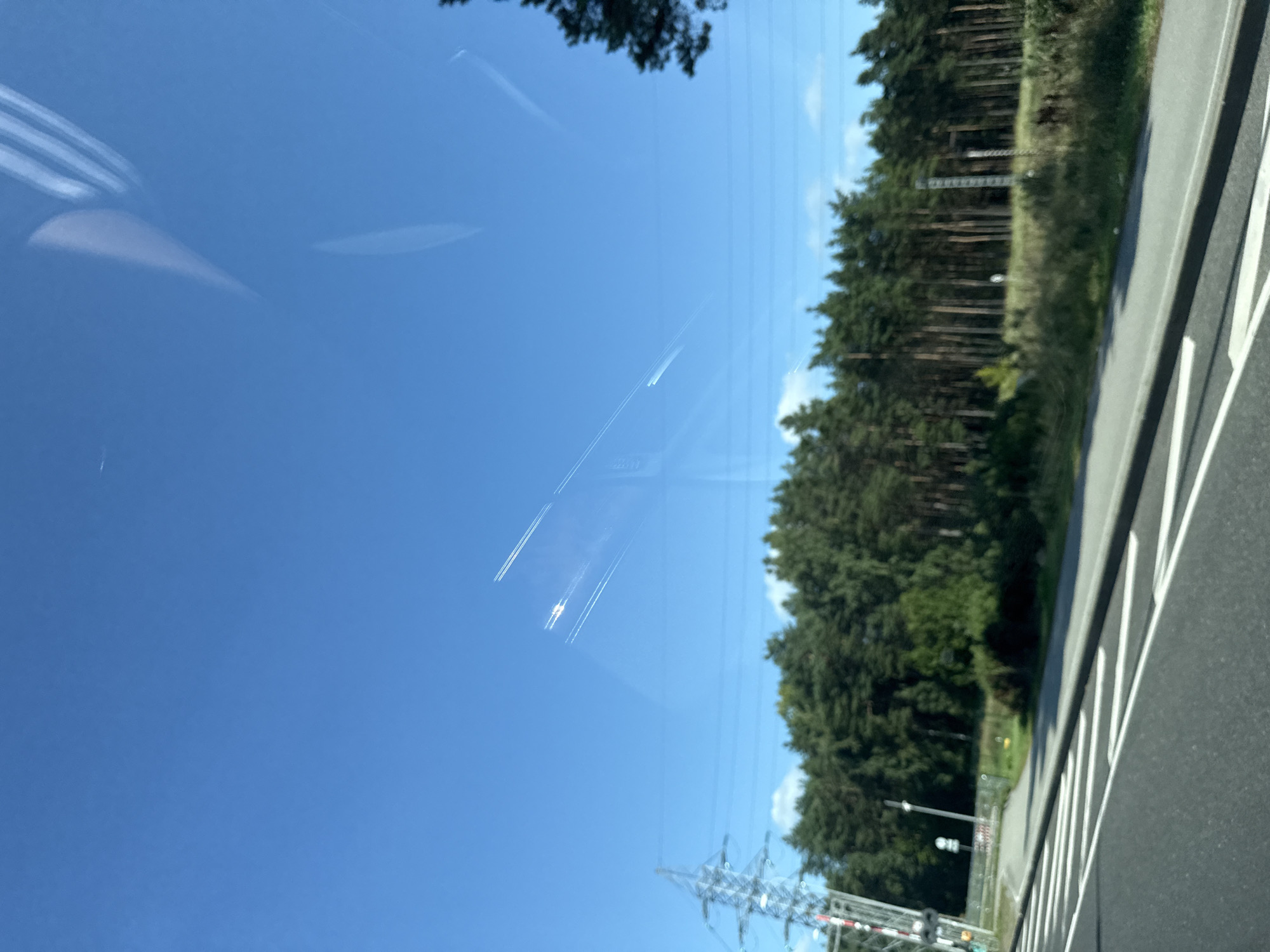 The image depicts a clear blue sky with several contrails from airplanes. In the foreground, a road with white markings is visible, suggesting a driving perspective. To the left, a large electricity pylon stands prominently. Beyond the road, a dense line of pine trees forms a forest, with some undergrowth and grass. The horizon is marked by the treeline and a bright, sunny sky. There are no people or animals visible in the image, and no specific landmarks that would definitively place the scene in Tallinn, Estonia. The overall impression is one of a rural or suburban landscape on a bright day.