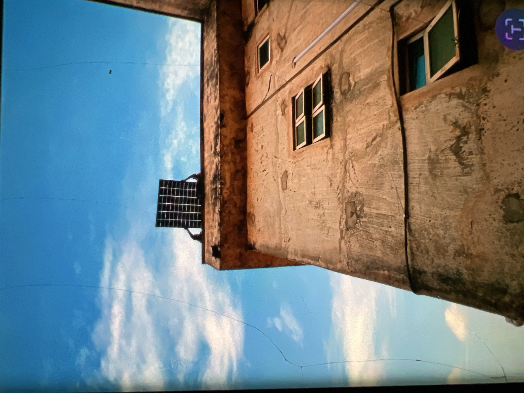 A low-angle, full shot captures a weathered, ochre-colored building against a bright blue sky dotted with white clouds, a scene likely set in Lagos, Nigeria, given its distinctive architectural style and the overall atmosphere. The building appears to be a multi-story structure, possibly a residential building or an older office block, characterized by its rough, textured facade and peeling paint.
On the rooftop, two hands are visible, holding a dark, rectangular solar panel. The hands appear to be positioned to install or adjust the panel. The solar panel itself is composed of many small, photovoltaic cells arranged in a grid pattern. The angle of the shot emphasizes the height of the building and the upward expanse of the sky, creating a sense of scale.
The building's exterior features several windows. On the visible wall, there are three windows. Two of these are arranged horizontally, one above the other, with green panes and wooden frames, both slightly ajar. Below these, another similar window is visible, also ajar. A third, smaller window is positioned higher up on the right side of the wall, with its frame and possibly a green pane, appearing closed. A thick, black cable runs horizontally across the building's facade, connecting to a vertical pipe that descends downwards. The texture of the concrete walls is visibly rough and uneven, showing signs of age and exposure to the elements. The overall impression is one of a functioning, albeit aged, urban structure in a tropical environment.