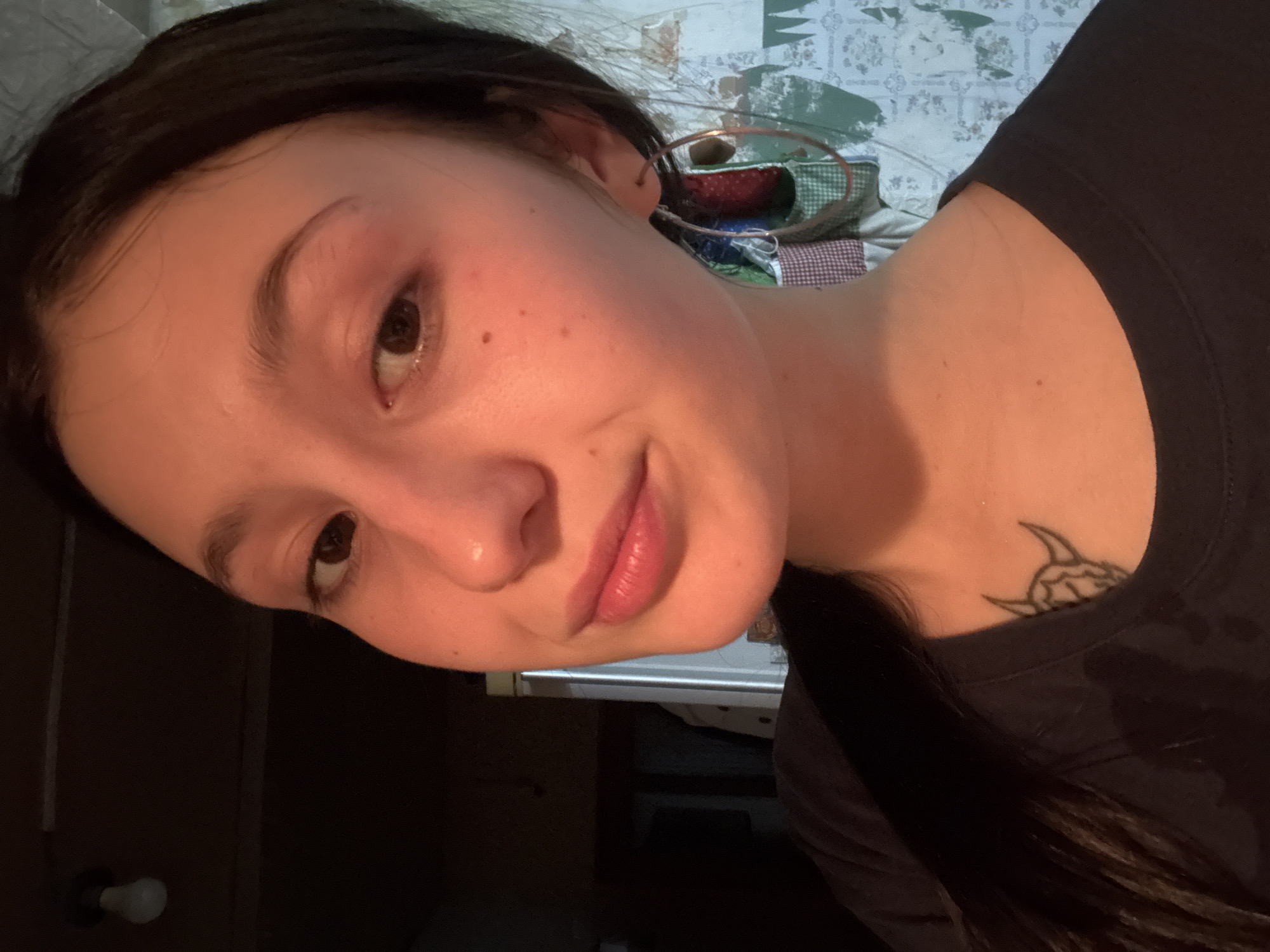 A close-up, tilted selfie of a young woman with dark hair. She has light skin, brown eyes, and is wearing minimal makeup. She is wearing a dark t-shirt, and a tattoo is visible on her chest/collarbone area. The background is blurry and indistinct, showing a bed and a wall with a patterned covering. The location in the city of Фоки, Russia is not discernible from this image.