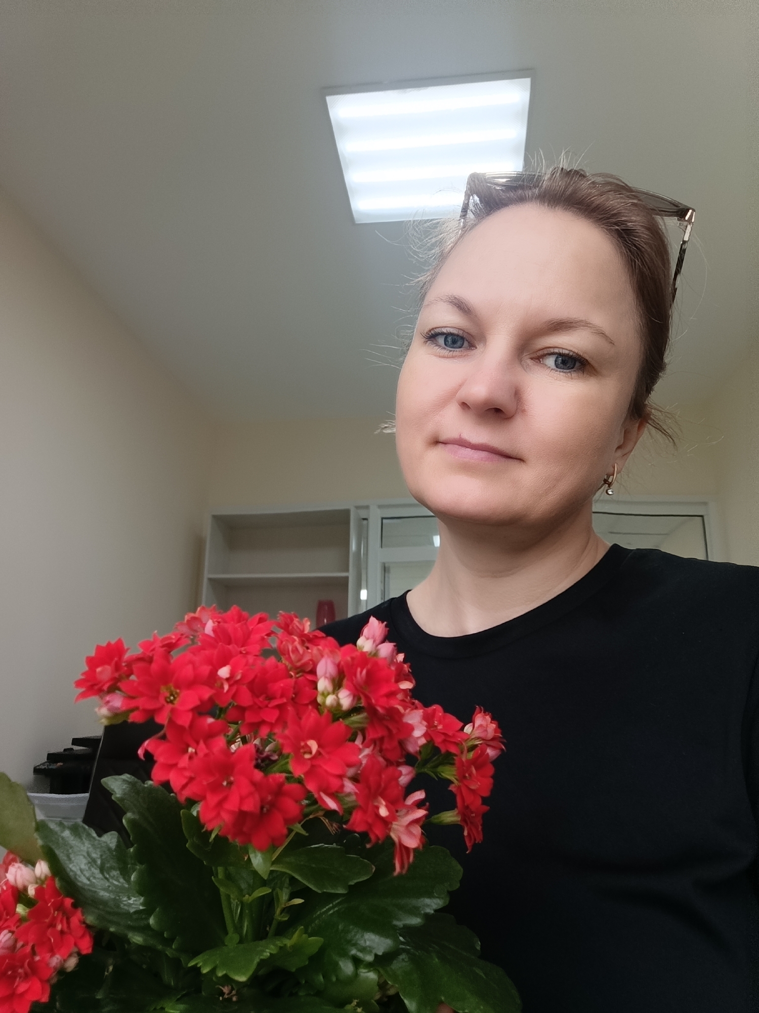 A woman is holding a red flowering plant. The woman has blue eyes, fair skin, and brown hair. She is wearing a black t-shirt and earrings. Her glasses are perched on her head. The plant has green leaves and clusters of small red flowers. The background is a room with beige walls, a white cabinet, and a fluorescent light fixture in the ceiling. The location is Марковский, Russia.