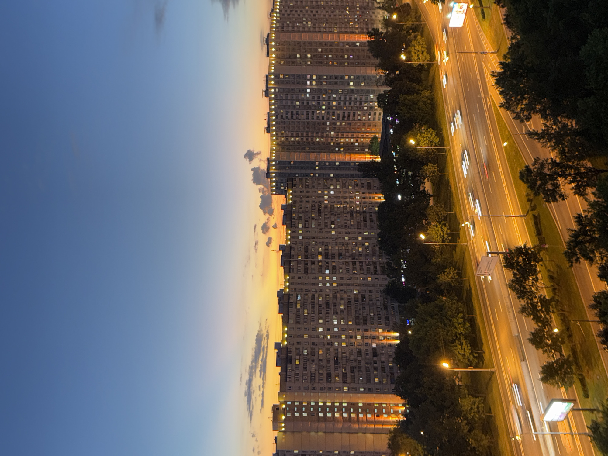 This is a high-angle, medium shot of a large, multi-story apartment building complex on the outskirts of Moscow, Russia, during twilight. The sky is a gradient of deep blue at the top, transitioning to a bright orange and yellow sunset glow near the horizon, with scattered clouds. The apartment buildings are long and rectangular, with numerous windows, many of which are illuminated, indicating residents are home. Decorative lights are visible along the rooftops and vertical accents of orange lighting illuminate sections of the building facades.
Below the buildings, a wide, multi-lane highway is visible, with cars depicted as streaks of light due to motion blur, suggesting active traffic. Streetlights cast a warm, yellow glow on the road and surrounding trees. The foreground features dense foliage and trees, obscuring the lower parts of the buildings and the immediate ground level. A digital billboard is visible on the right side of the highway, illuminated with white light. The overall scene depicts a typical urban environment in Moscow as night begins to fall, with the juxtaposition of residential buildings and active infrastructure.