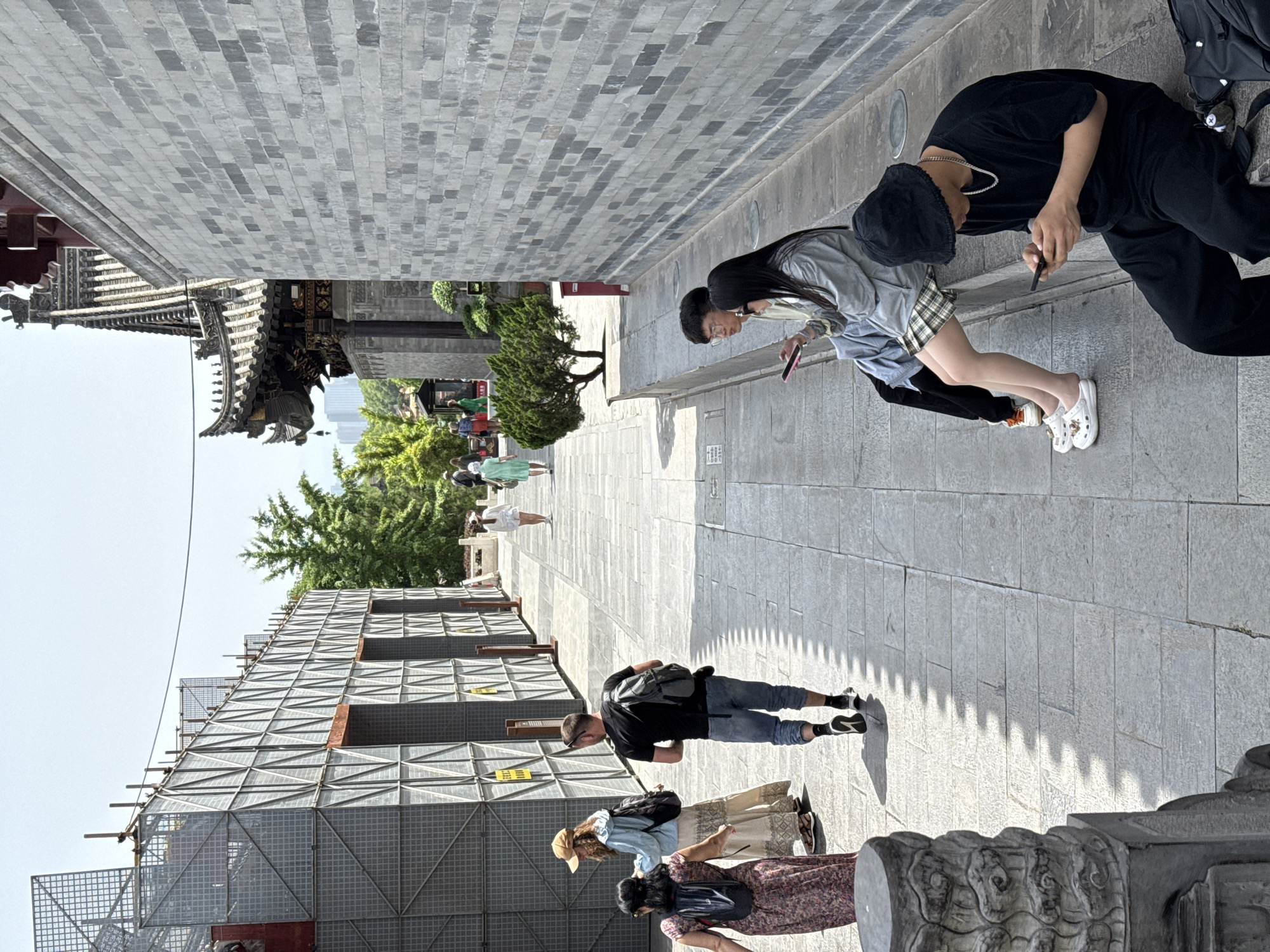 The photo shows a street scene in the Yanta District of China, defined by a narrow pathway flanked by buildings and walls made of gray bricks. A couple in the foreground are looking at their phones. The man is wearing a black outfit and the woman a plaid skirt and gray sweater. Behind them and further into the distance, other people are walking along the path. On the left, a building is covered with scaffolding. The sky is light.