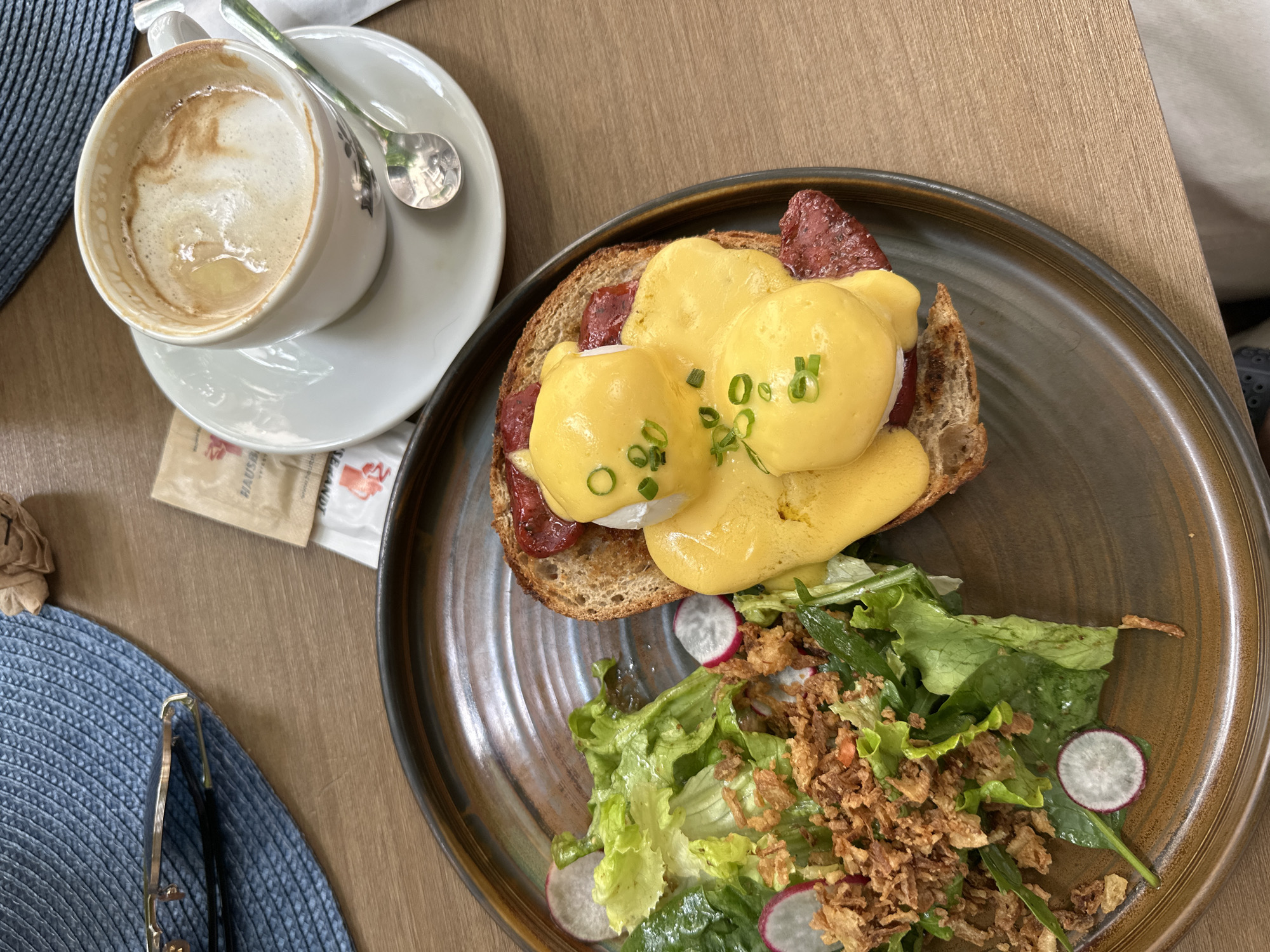 The image displays a breakfast setting on a wooden table. A plate contains Eggs Benedict with hollandaise sauce, served on toast with what appears to be bacon. A side salad with lettuce, radishes, and crispy fried onions is also present. A cup of coffee with latte art and a spoon rests on a saucer. A pair of sunglasses and a sugar packet are visible on a blue woven placemat. The setting suggests a cafe or restaurant in Sofia, Bulgaria, given the typical offerings and ambiance.