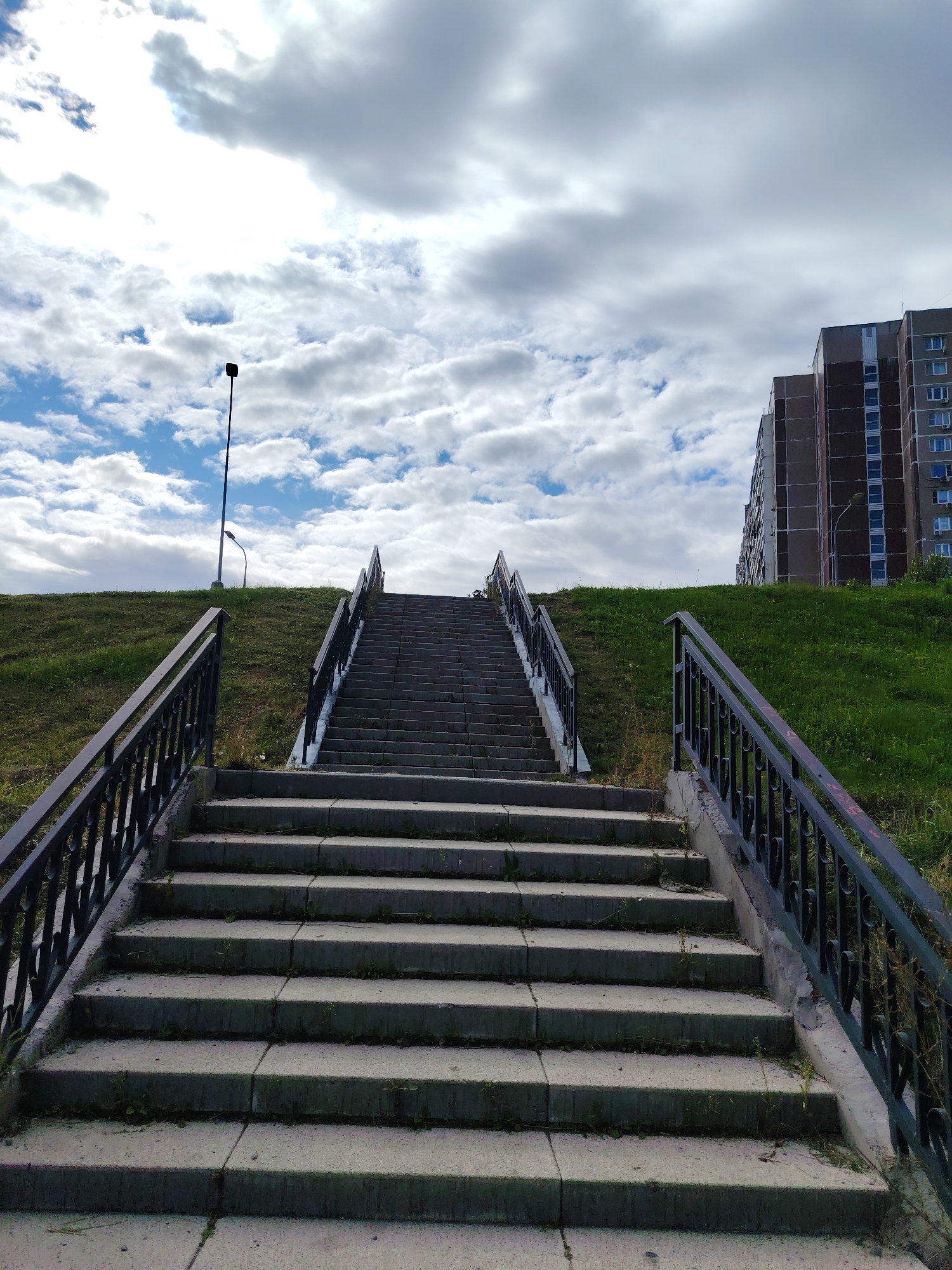 A wide, outdoor, low-angle, eye-level shot captures a concrete staircase with black metal railings leading up a grassy hill under a partly cloudy sky. To the right of the staircase and hill, a tall, multi-story residential building with a brown facade and numerous windows is visible. A single streetlamp on a thin pole stands on the left side of the hill. The scene is devoid of people and exhibits a typical urban environment. This scene is located in the city of Zelenograd, Russia.