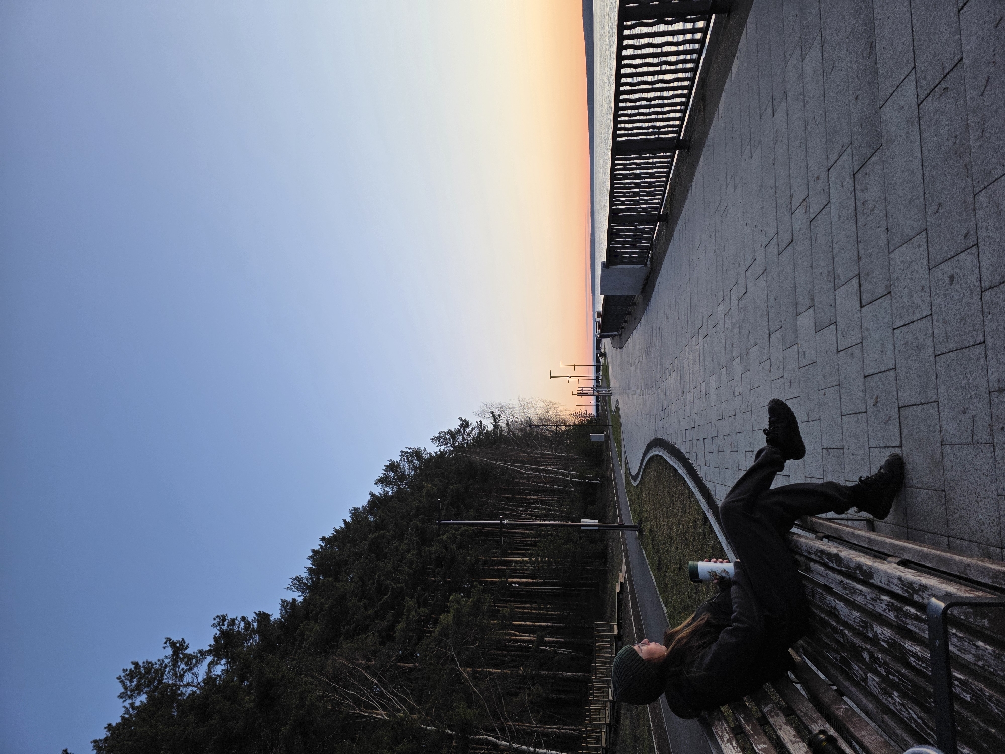 A serene outdoor scene at dusk or dawn captures an individual resting on a bench along an embankment in Chaikovsky, Russia, overlooking a body of water and a forest.
In the foreground on the right, a person, dressed in a dark jacket, pants, and a beanie, lies sideways on a wooden slatted bench. They hold a white and green thermos cup in their hand, appearing relaxed and gazing towards the horizon. Their legs are bent, with their black boots resting on the back of the bench.
To the person's right, a grey paved promenade or path stretches into the distance, gently curving. A dark metal railing separates the path from a vast, calm body of water, likely a river or reservoir, which reflects the soft light of the sky. To the person's left, a narrow strip of grass borders the path, followed by a dense forest of dark evergreen and some bare deciduous trees situated on a sloping bank. Several tall streetlights are visible along the path and amidst the trees.
The background features a clear sky displaying a beautiful gradient of colors, transitioning from soft peach and orange hues near the horizon to a clear, pale blue overhead. This coloration, combined with the subdued lighting, strongly suggests either sunset or sunrise. The overall atmosphere is peaceful and contemplative. No discernible text is visible on the thermos or elsewhere in the image.