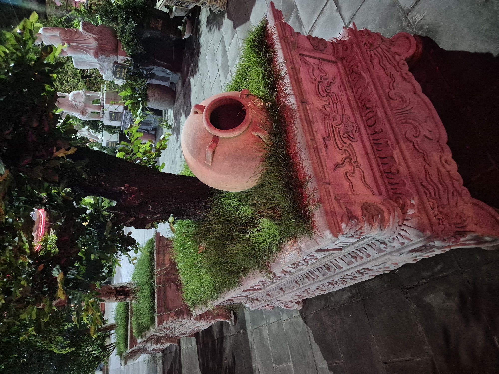 The image captures a serene nighttime scene in Đà Nẵng, Vietnam. In the foreground, a large, ornate, stone planter, bathed in a reddish light, contains lush green grass and the base of a tree. A large, terracotta-colored amphora is tilted on its side within the planter, partially submerged in the grass, with its opening facing upwards. The planter itself features intricate carvings and rests on decorative feet, with a row of similar planters receding into the background. Beyond the planters, white marble statues of figures, including one on an elephant, are visible, adding a cultural and artistic element to the scene. The ground is paved with dark square tiles, reflecting the surrounding light. The overall impression is one of peaceful contemplation and cultural heritage, likely within a temple or garden complex.