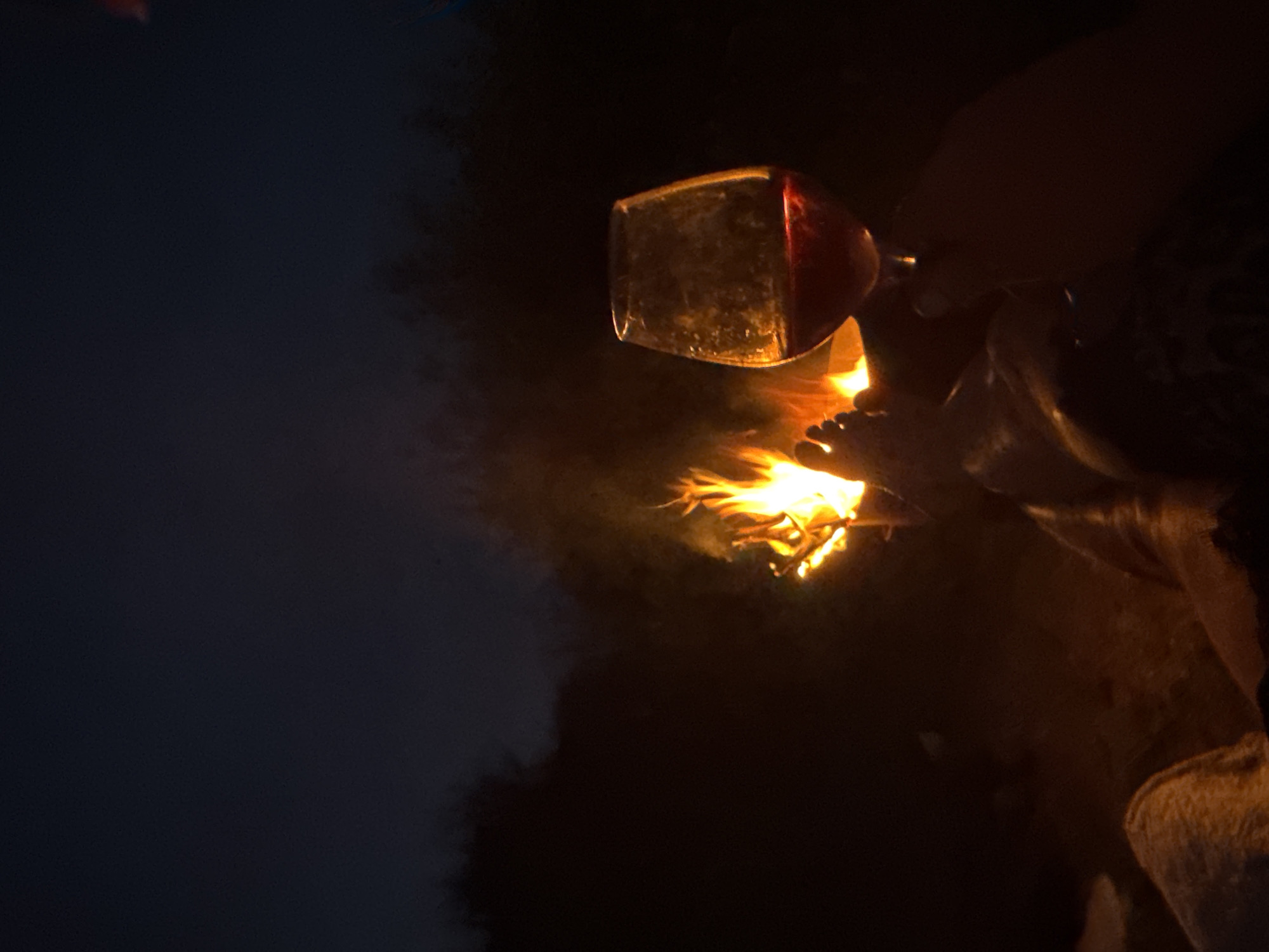 In the dim light of a campfire, a person's feet are visible in the foreground, positioned near the flames. A hand holds a stemmed glass filled with a red beverage, likely wine, up to the right. The background is dark, with the silhouette of trees against the night sky, suggesting an outdoor setting. The image evokes a sense of relaxation and warmth. The location is not specified in the image or the provided crops.