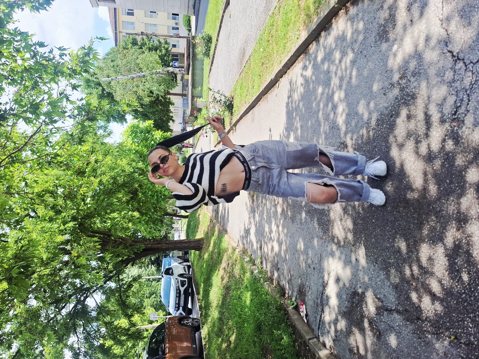A young person stands on a cracked asphalt path, posing for a photograph in a sunny outdoor setting in Mezdra, Bulgaria. The individual, likely female, has dark hair pulled into a high ponytail and wears dark oval sunglasses. She is dressed in a black and white striped cropped sweater, light-wash ripped jeans that reveal her knees, and white sneakers. A "PULL&BEAR" branded waistband is visible above her jeans, and she has an unidentifiable text tattoo on her midriff. Her left hand is raised near her face, while her right hand lightly tugs at her ponytail.
To her left, behind a grassy verge, are several parked vehicles, including a white SUV with the Bulgarian license plate "BH 6339 CH" and a brown SUV/truck. Lush green trees provide shade, casting dappled light and shadows across the path. In the background, multi-story residential buildings are visible, alongside more dense greenery. A blue pedestrian crossing sign is seen in the middle distance near what appears to be a narrow body of water or canal. To her right, the paved path continues alongside a grassy strip containing some small rose bushes. A discarded red and white item of trash lies on the ground near the curb. The strong sunlight and pronounced shadows indicate it is midday or early afternoon.