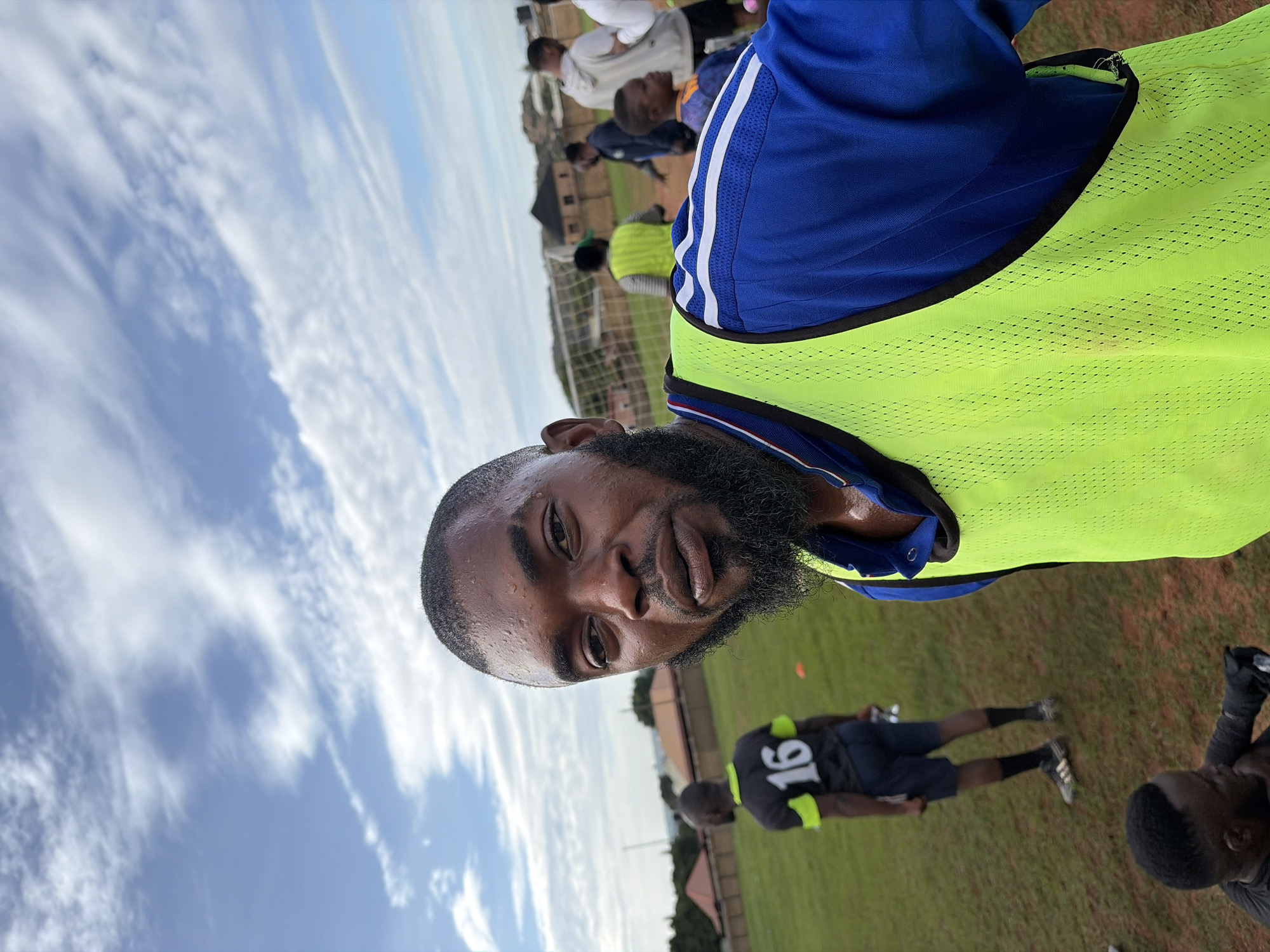 The image presents an outdoor scene on a sports field in Jos, Nigeria, under a bright blue sky with scattered white clouds. The primary subject, a dark-skinned man with a short haircut and a full beard, occupies the foreground, looking directly at the camera. He is visibly perspiring, with sweat visible on his forehead and upper lip. He wears a blue long-sleeved athletic shirt featuring white shoulder stripes, overlaid with a bright neon green mesh training bib.
In the mid-ground to the left, another dark-skinned individual is observed from the back, wearing a black or dark grey t-shirt with neon green trim and the white number "16" on the back. This person wears dark shorts and athletic shoes and appears to be holding a clear plastic bottle. An orange training cone is situated on the field behind this individual.
To the right, a white soccer goal net is visible. Multiple other individuals are dispersed across the background of the field, including a person near the goal wearing a neon yellow training bib, a person in a white hoodie, another in a dark long-sleeved shirt, and a small child in a blue and orange shirt. The field itself is covered in grass, interspersed with visible patches of dry, reddish-brown earth. Beyond the field, several single-story buildings with reddish-brown and dark roofs are discernible, indicating a residential or semi-urban environment. The top of a person's head and shoulder are partially visible in the bottom-left corner of the foreground.