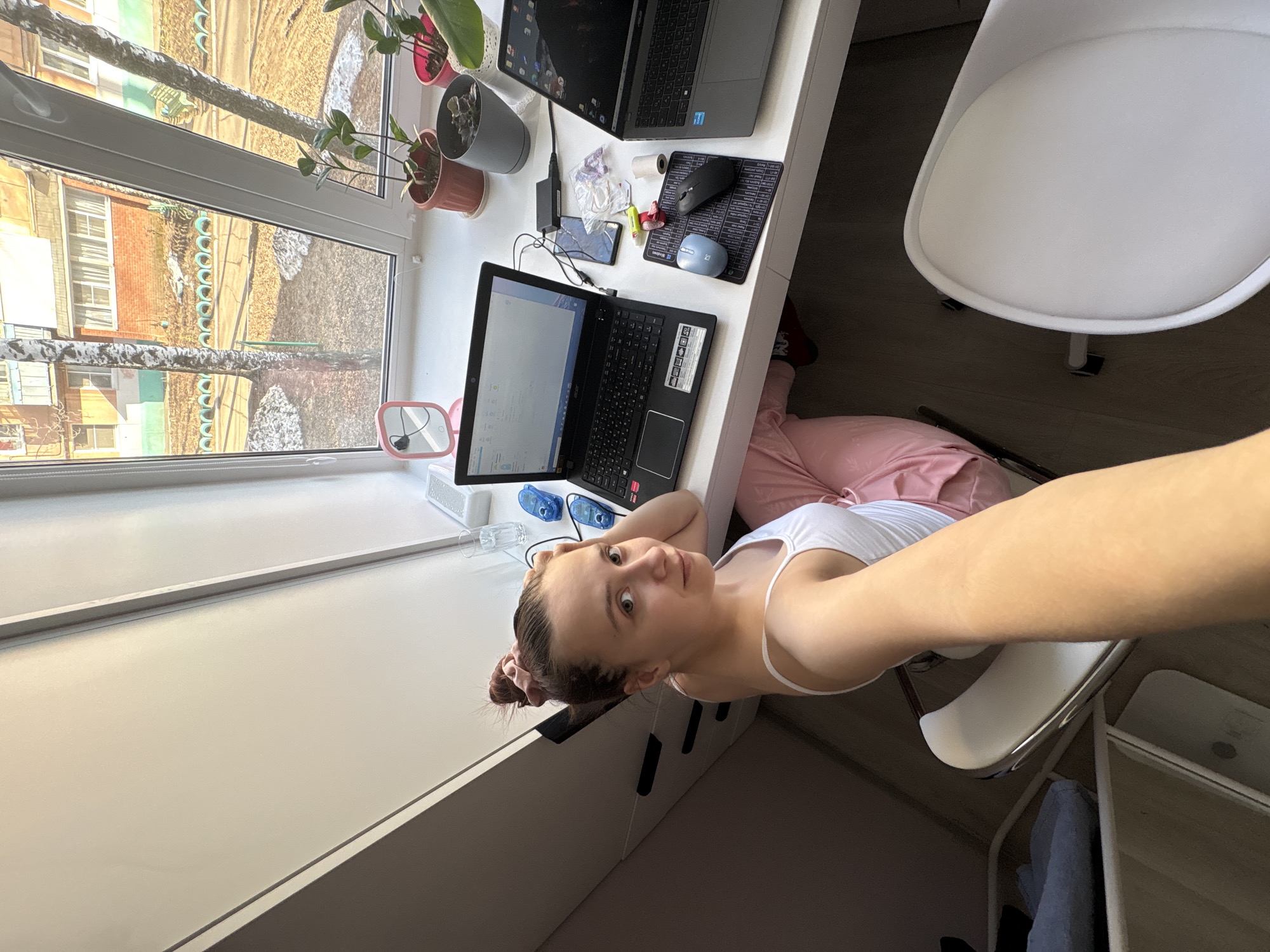 A woman with her hair pulled back in a bun is sitting in a white chair in front of a white desk in what appears to be an indoor space. The desk has two laptops, a mousepad, a phone, and various office supplies. Outside the window, a birch tree and building are visible, suggesting the location is in a residential area. Based on the appearance of the buildings and environment, the location is the city of Chaikovsky, Russia. The woman is taking a selfie. She is wearing a white tank top and pink sweatpants.
