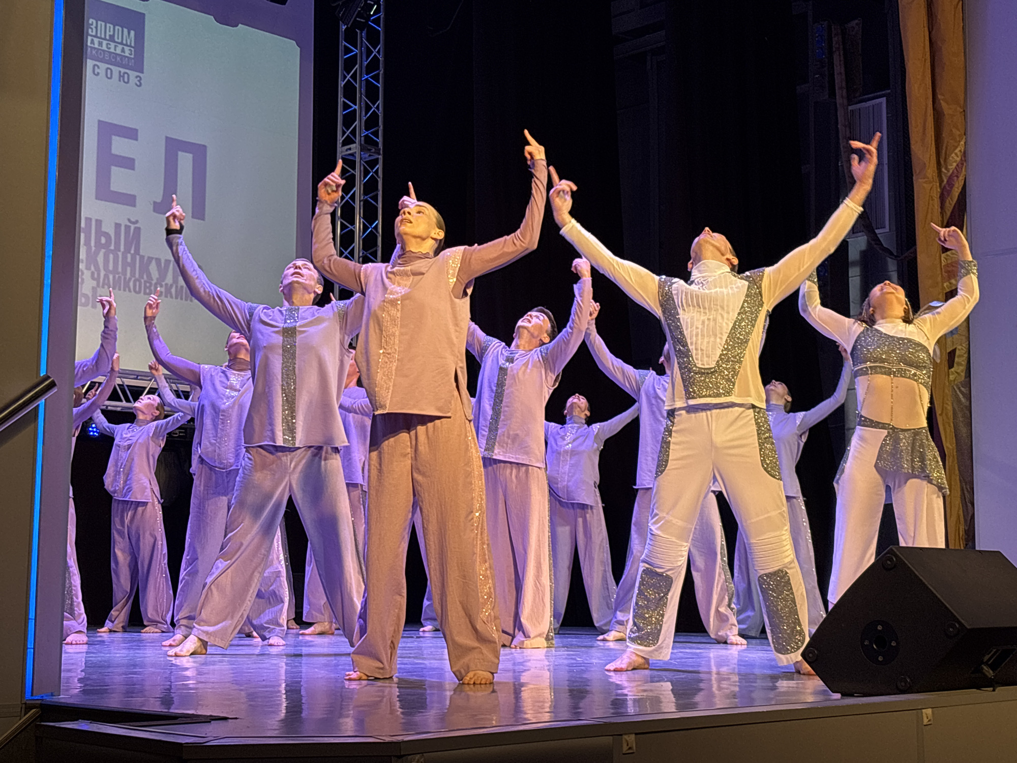 A dance performance is taking place on a stage. The stage is lit with purple lighting and has a glossy floor. At least ten dancers are present, dressed in varying outfits of similar color tones. Some wear pale purple pajama-like sets with silver accents, while others are in white and silver ensembles. All dancers are barefoot, their arms raised and fingers pointing upwards. A large screen is visible in the background, displaying text in a non-English script, with "ЧАЙКОВСКИЙ" (Chaikovsky) at the bottom, which indicates the location in the city of Chaikovsky, Russia. Additional stage equipment, including speakers, are visible at the front of the stage. A vertical stack of metal bars is visible on the left-hand side.