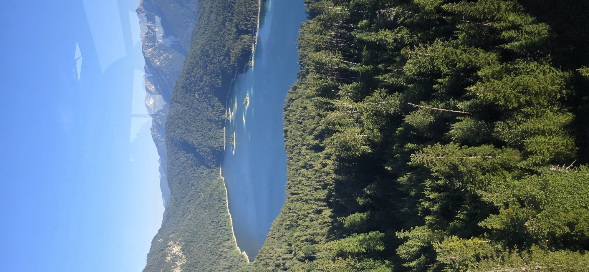 A high-angle shot captures a serene natural landscape with a clear blue sky and a large, tranquil lake surrounded by lush green forests and majestic mountains. The scene is Grainau, Germany. The lake is a vibrant blue, reflecting the sky, and several small, tree-covered islands dot its surface. The dense evergreen forest blankets the rolling hills that lead up to the mountains in the distance. The mountains themselves are rugged and rocky, with some appearing snow-capped. The lighting suggests it is daytime, possibly morning or afternoon, with soft sunlight illuminating the scene. There are no people or man-made structures visible in the immediate foreground or within the lake area, emphasizing the pristine natural beauty of the location. There are some reflections visible on what appears to be a window pane, which is likely where the photo was taken from.