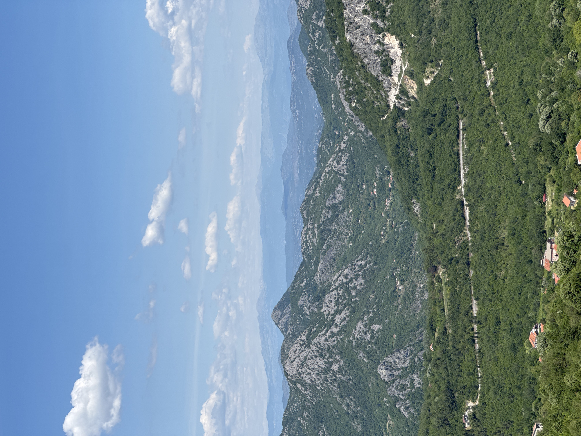 The image shows a mountainous landscape in Bukovik, Montenegro. The sky is blue with scattered white clouds. Multiple layers of mountains are visible, fading into the distance. The closest mountains are covered with dense green vegetation and rocky outcrops. A road winds through the vegetation, and houses can be seen near the edge.