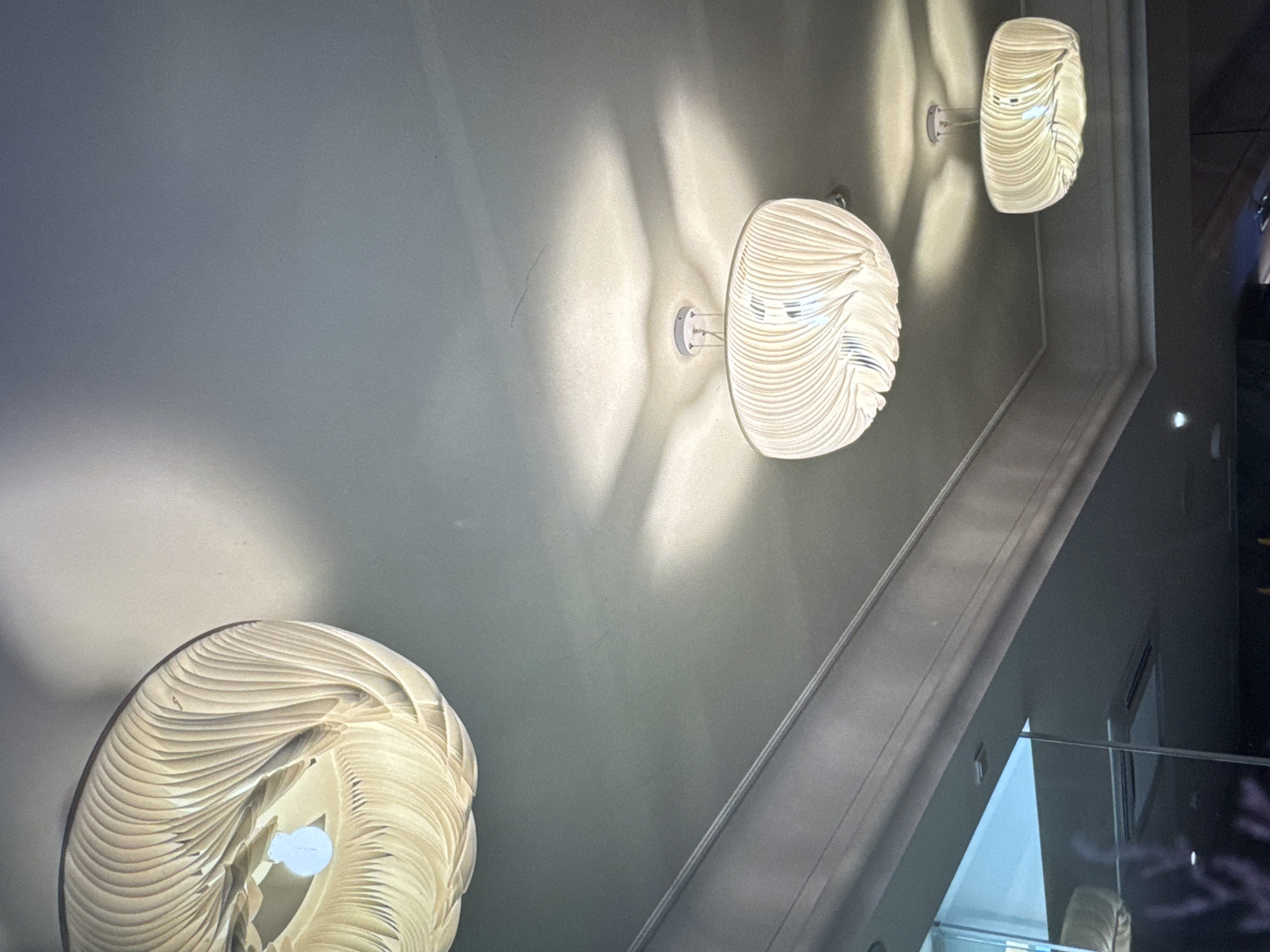 The image shows three decorative, white, circular wall lamps in a descending line. They are illuminated and casting a soft light on a gray ceiling. The bottom lamp is most prominent. A segment of what appears to be a glass partition or balcony railing is visible in the bottom right corner. Based on the location data provided, it is likely that the scene is located in Новинки, Russia.
