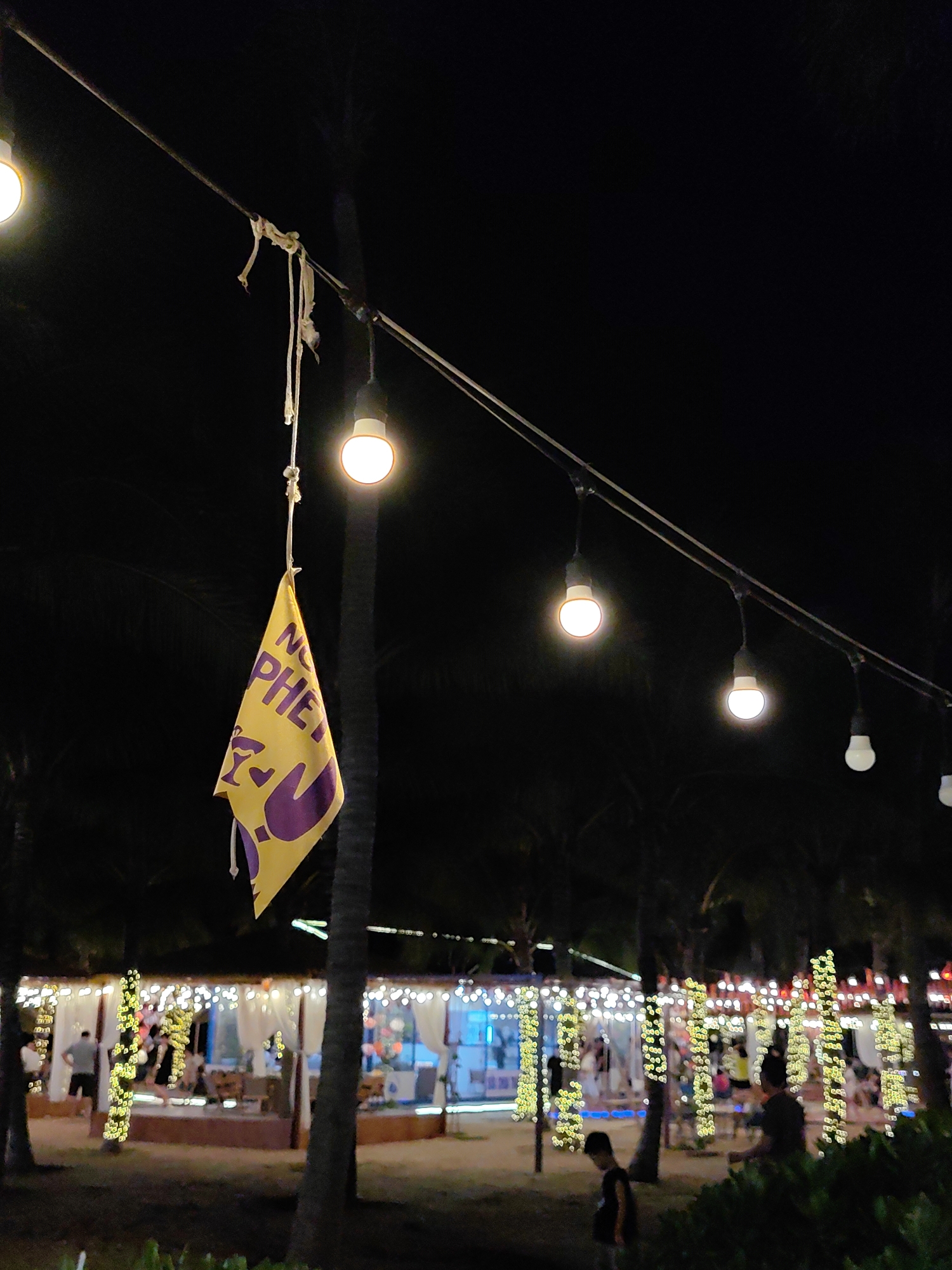 The scene is an outdoor evening view in Bai Chay Ward, Vietnam, illuminated by strung lights. In the foreground, a yellow flag with purple lettering and symbols is attached to the string of lights by a rope. Several lit light bulbs hang from the same line. In the midground, partially obscured by palm trees, are structures decorated with string lights and white curtains. People are visible inside these structures, possibly socializing or dining. A young boy stands on a walkway in the lower foreground, while other figures are seen at the edges of the frame. The overall environment appears to be a beachside or resort setting. The sky is dark, suggesting it is nighttime.
