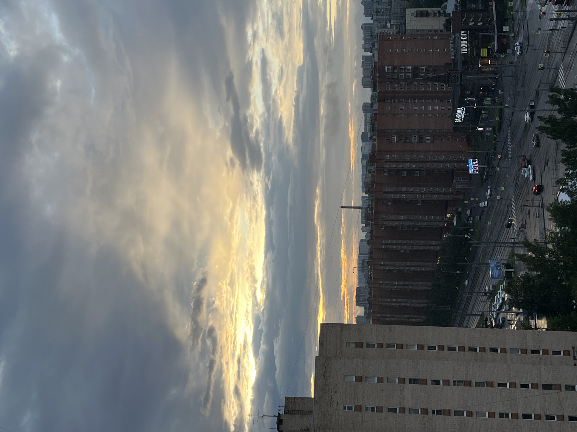 The image depicts an urban cityscape under a dramatic sky, likely at dusk or dawn. In the foreground, a tall, light-brown brick building with numerous rectangular windows occupies the left side of the frame. To its right, a long, dark red brick apartment building stretches across the mid-ground. A street with traffic lights, street lamps, and parked cars is visible in front of this building. Further in the distance, a dense cluster of multi-story buildings forms the city skyline, fading into a hazy background. Above the city, the sky is filled with heavy, dark gray clouds interspersed with streaks of bright yellow and orange light, suggesting the sun is low on the horizon. A few tall, thin structures, possibly chimneys or antennas, are visible among the buildings. The overall impression is one of an overcast, active urban environment.