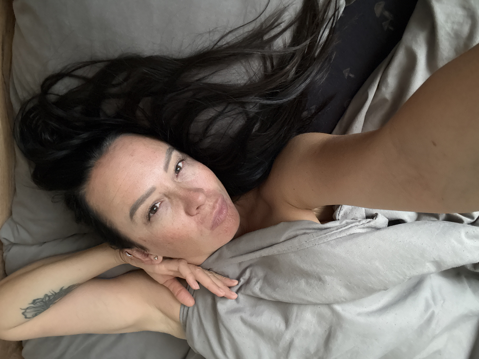 A young woman with long, dark hair is lying on a bed, looking at the camera. She is partially covered by a grey blanket, with her arm resting on her forehead. The room appears to be a bedroom, with grey sheets and pillows. There is a tattoo of a feather on her upper arm. The image was taken in Chaikovsky, Russia.
