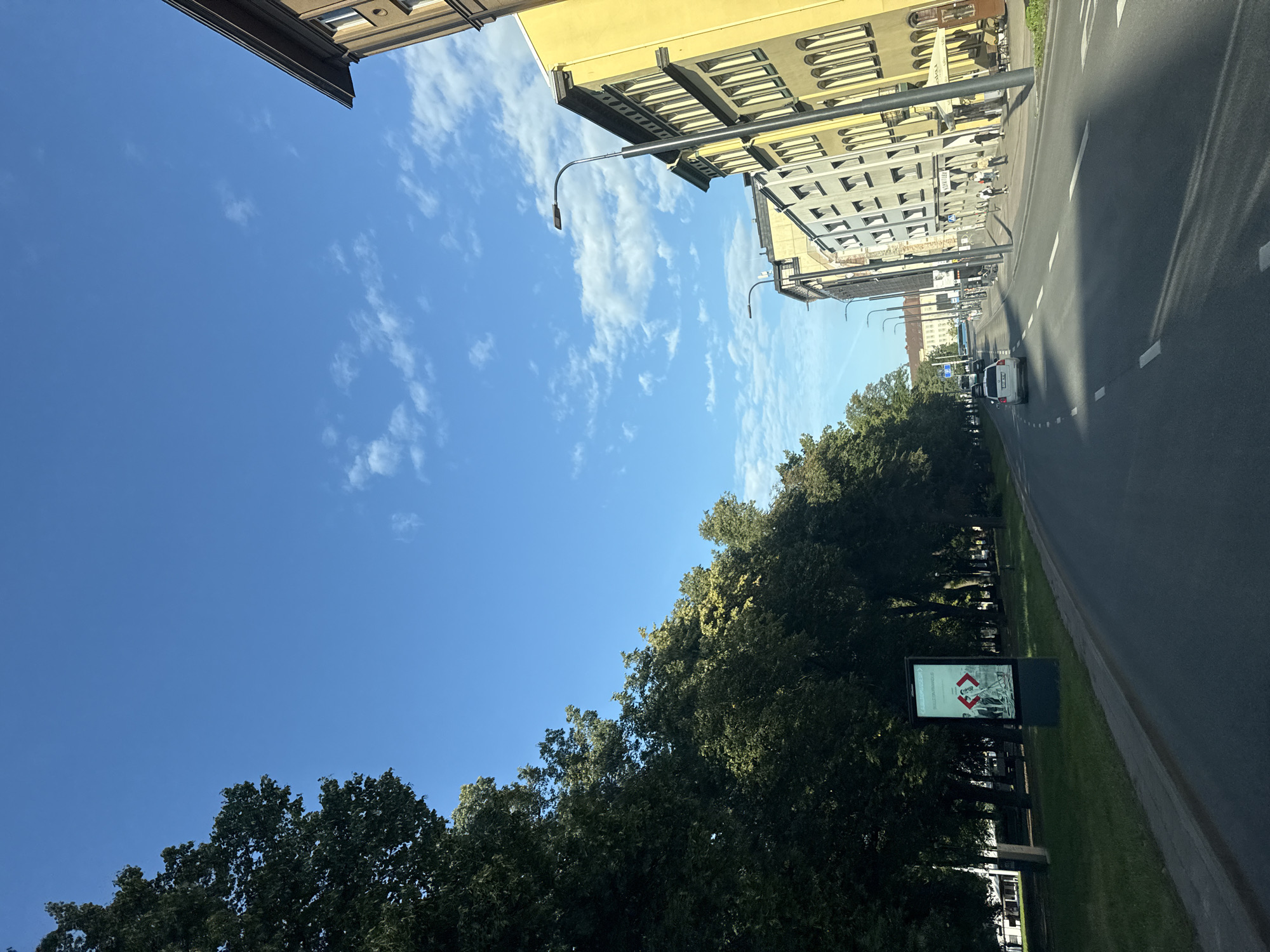 The image shows a street scene in Tallinn, Estonia, looking down a road lined with trees on the left and buildings on the right. The sky is blue with scattered white clouds. On the left side of the road, there is a grassy area with a tall billboard. Further down the road, trees form a dense line. On the right side, there are several multi-story buildings with a predominantly yellow facade on one of them, featuring many windows. Pedestrians are visible walking on the sidewalk. A white car is driving on the road. Streetlights are present along the right side of the road. The road surface is asphalt with white lane markings. The overall scene depicts a typical urban environment in Tallinn on a sunny day.