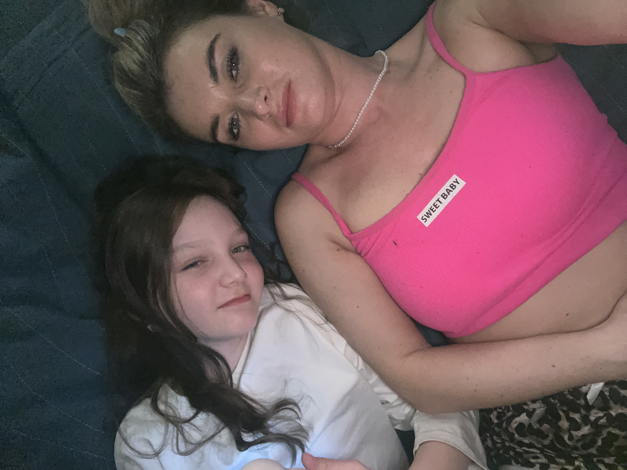 Two individuals, an adult female and a child, are lying down indoors. The adult female is positioned at the top of the frame, wearing a pink tank top with a "SWEET BABY" label on it and a pearl necklace. She has her hair styled in a bun. The child lies beneath her, wearing a white top. The setting appears to be on a blue quilted surface. The location is not apparent from the image and no information suggests it is specifically in the city of Chaikovsky, Russia.