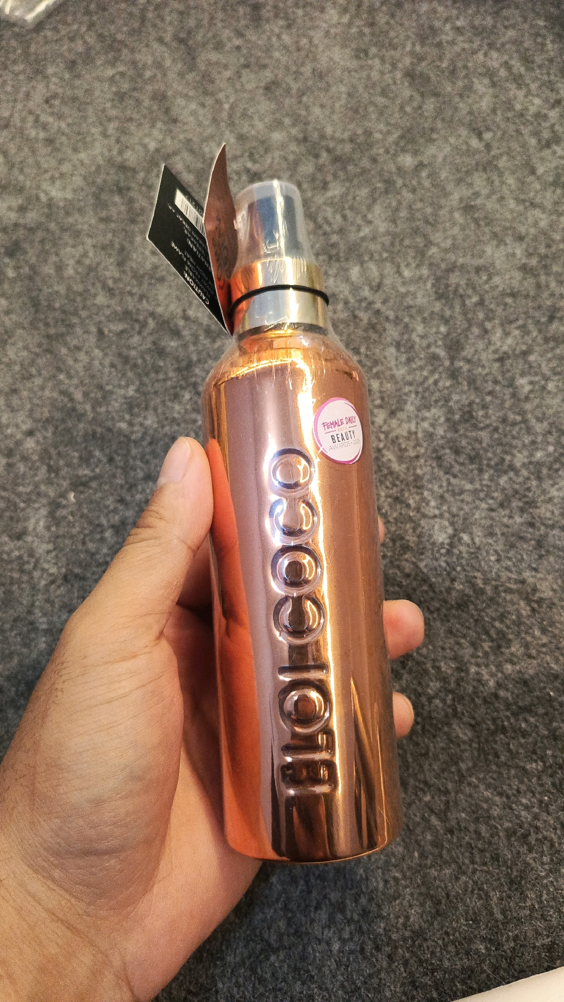 A hand is holding a rose gold colored spray bottle with the word "COCO" embossed on it. A pink sticker with the text "FEMALE DAILY BEAUTY AWARDS 2020" is on the bottle. A black tag is attached to the bottle. The background is a grey textured surface, possibly carpet or fabric. There is no indication of the location being in Babadan, Indonesia.