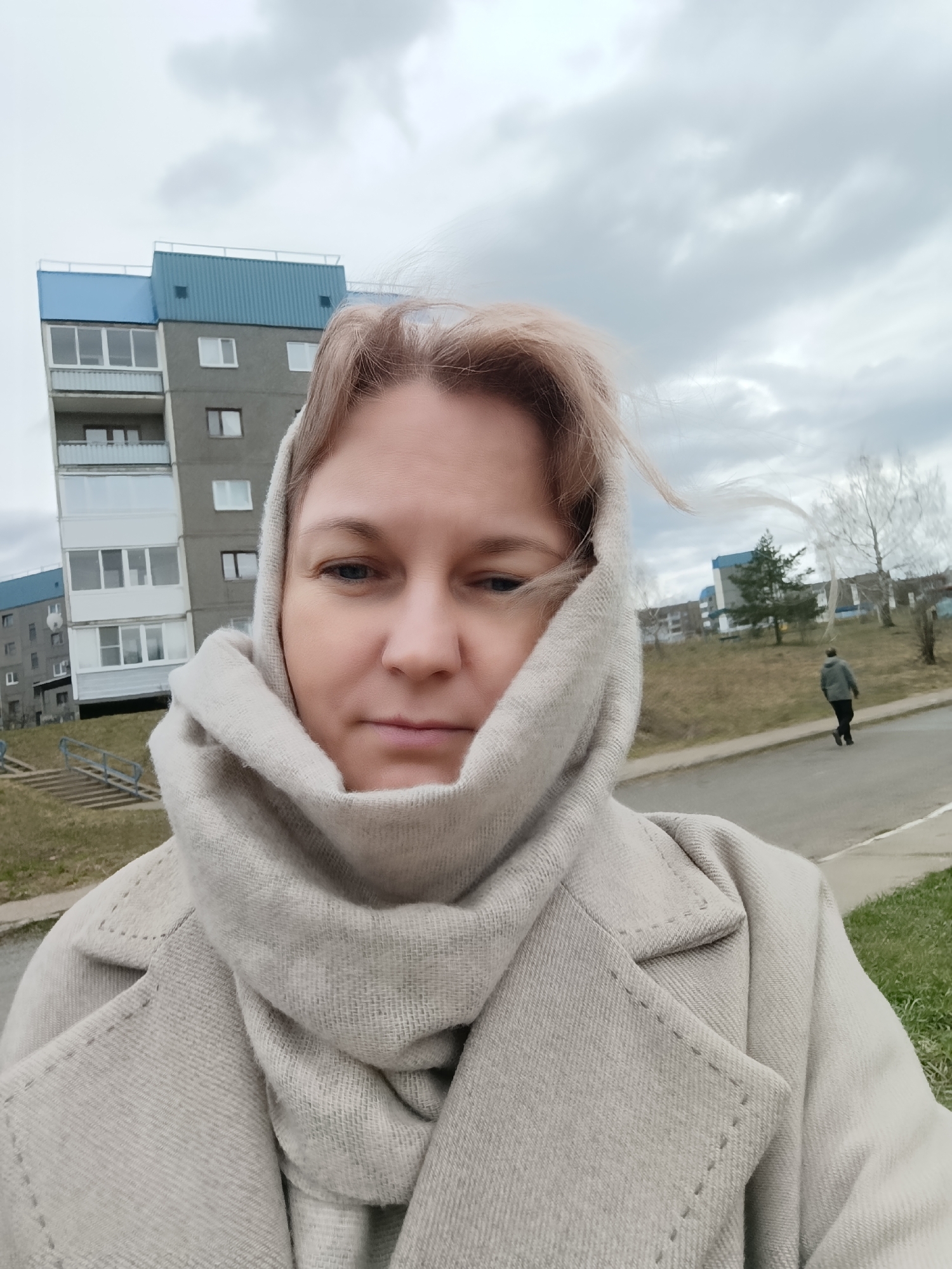 A medium-shot photograph shows a woman wearing a light-colored coat and scarf wrapped around her head. She faces the camera with a neutral expression. In the background is a multi-story building with blue accents, typical of urban architecture. A person walks along a sidewalk adjacent to a patch of grass. The sky is overcast, suggesting a cold or temperate climate. The scene is set in the city of Марковский, Russia.