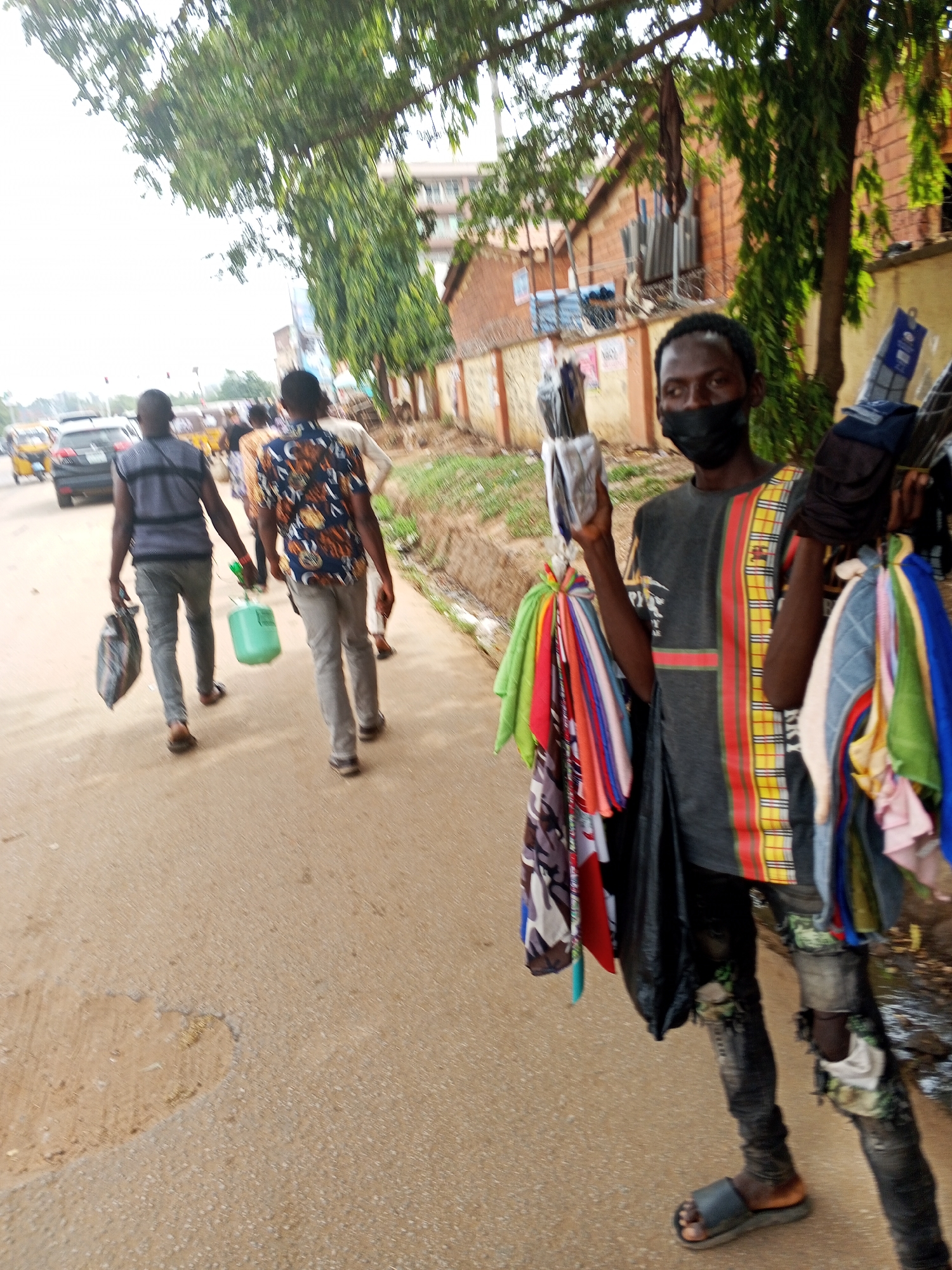 A daytime street scene in Abuja, Nigeria shows a male vendor wearing a black face mask, a gray t-shirt with a vertical red and yellow striped design, torn jeans, and flip-flops. He holds up folded clothes in his left hand and carries more items on a pole over his shoulder, including scarves and other textiles. Behind him, two men walk away on a paved road. One wears a vest and jeans, carrying a bag, and the other wears a patterned shirt and jeans. A light-colored car is visible in the distance. The background includes buildings, trees, and a wall. The weather is overcast.