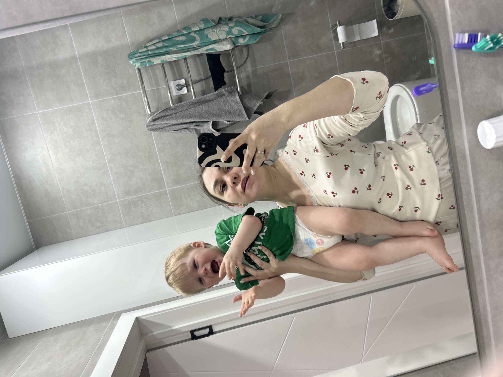 A mirror selfie, rotated 90 degrees clockwise, captures a woman holding a young child in a modern bathroom.
The woman, with dark hair, is wearing a cream-colored long-sleeved top adorned with a red cherry pattern. She holds a black smartphone with a distinctive black and white swirly-patterned case in her right hand, taking the picture. A simple gold ring is visible on her left hand. Her expression is neutral to slightly smiling.
She is holding a blonde-haired toddler, who is wearing a bright green t-shirt and a disposable diaper with the number '4' clearly visible on it. The child's mouth is wide open, suggesting crying or a loud vocalization.
The bathroom features light gray rectangular tiles on both the walls and floor. In the reflection, a white toilet is visible in the background, along with a chrome toilet paper holder holding a roll of paper. Above the toilet, a metal towel rack holds a folded teal patterned towel and a gray towel. To the right, various colored toothbrushes (purple, green, blue) are mounted on the wall. Below the child's legs, a long white, minimalist cabinet or bath panel is visible. The scene suggests an indoor, well-lit environment, likely a home in Chaikovsky, Russia.