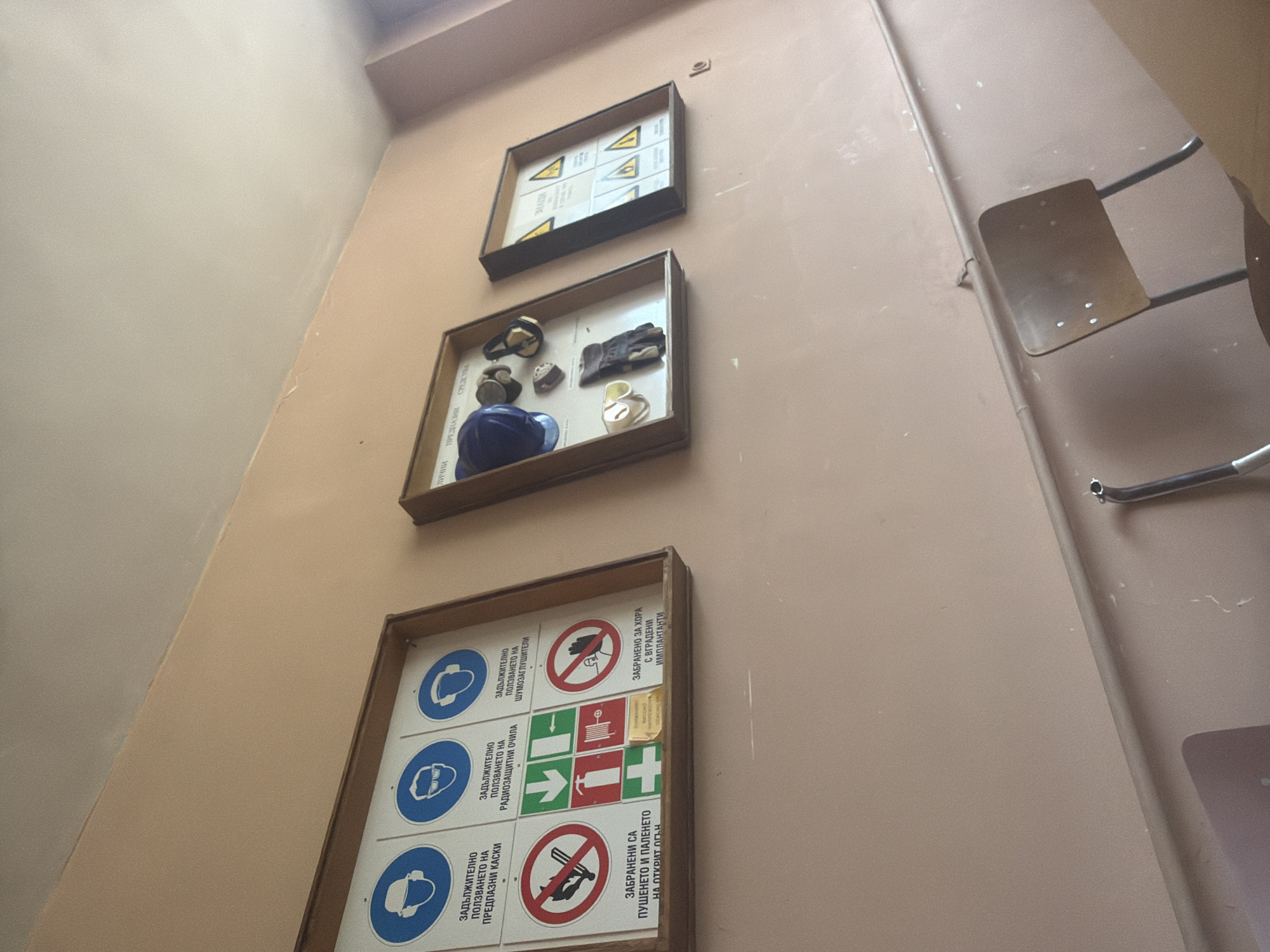 The image shows a wall in what appears to be a building, likely in Dolna Mitropolia, Bulgaria, as indicated by the Bulgarian text on the signs. Three framed displays hang vertically on the wall. The top one features hazard warning signs. The middle display contains a variety of safety equipment, including a hard hat, goggles, gloves, and a respirator. The bottom display is a grid of safety regulation signs, displaying various "mandatory use" and "prohibited" symbols related to hearing protection, eye protection, hard hats, smoking, and hazards for people with implants. To the right of the displays, a metal folding chair is partially visible, mounted on the wall. The wall is a muted orange color.