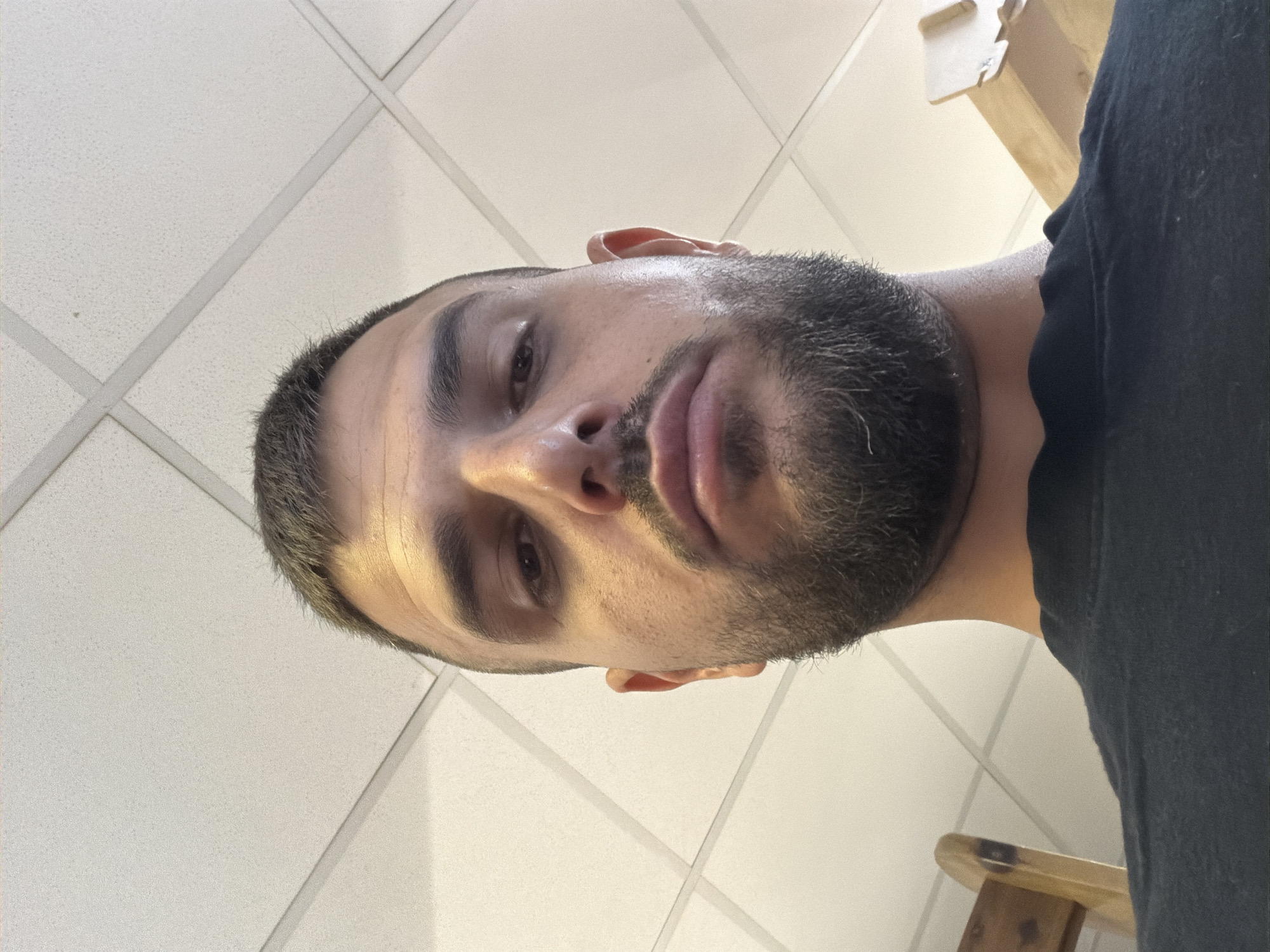 An inverted close-up portrait shows a man looking towards the camera from a low-angle perspective. The man has a short, well-trimmed dark beard and mustache, with short dark hair on his head. He wears a plain dark (likely black) t-shirt. His expression is neutral and his lips are slightly parted. The lighting appears to be artificial, originating from above, casting subtle shadows on his face, particularly beneath his brow and chin. His skin has a slightly reflective or oily appearance, notably on his forehead and nose. A small, light-colored fiber or lint is visible on the hair near his left temple. In the background, a light-colored, possibly off-white, drop ceiling with a grid pattern is visible, indicating an indoor setting. A wooden object, possibly part of a chair or furniture, is partially visible in the upper and lower right corners of the image. There is no visible text. The image likely depicts an indoor environment in Sofia, Bulgaria.