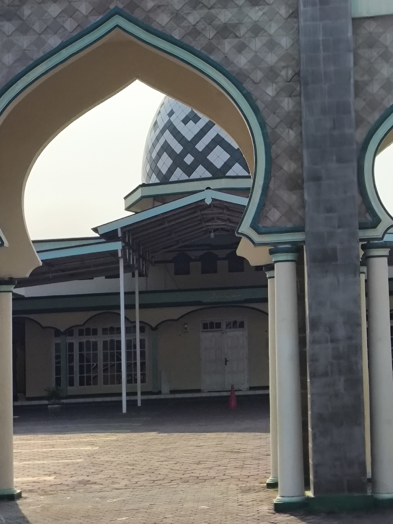 A mosque with a diamond-patterned dome and a light yellow facade is visible through a large archway with a pale yellow and teal trim. The archway is framed by two large stone pillars, one with a rough grey stone finish and the other partially visible and lighter in color. A tiled courtyard with a brick-like pattern stretches out in front of the mosque. The mosque features a covered walkway supported by white poles in front of several large windows and a double door. A small red traffic cone is visible on the courtyard. The sky is overcast. The image appears to be taken in Cilegon, Indonesia, likely in the vicinity of a mosque based on the architectural features. No people are present in the frame.