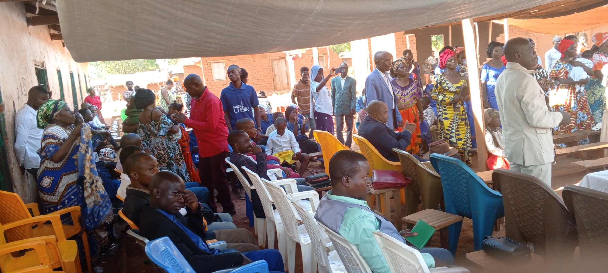 A wide-angle shot captures a gathering of people in what appears to be an outdoor or semi-outdoor setting in Kalemie, Democratic Republic of the Congo.
A group of people are seated in rows of white plastic chairs under a makeshift canopy, while others stand around the perimeter. The seated individuals, predominantly men, are dressed in casual attire, including shirts, vests, and suits. Some are engaged in conversation, while others appear to be listening or observing.
In the background, a group of people are standing, many of whom are women dressed in colorful, patterned clothing. They seem to be interacting or watching the proceedings.
The backdrop consists of buildings constructed with reddish-brown bricks or mud, typical of the region. The setting is simple and functional, suggesting a community event or meeting taking place in a residential area.