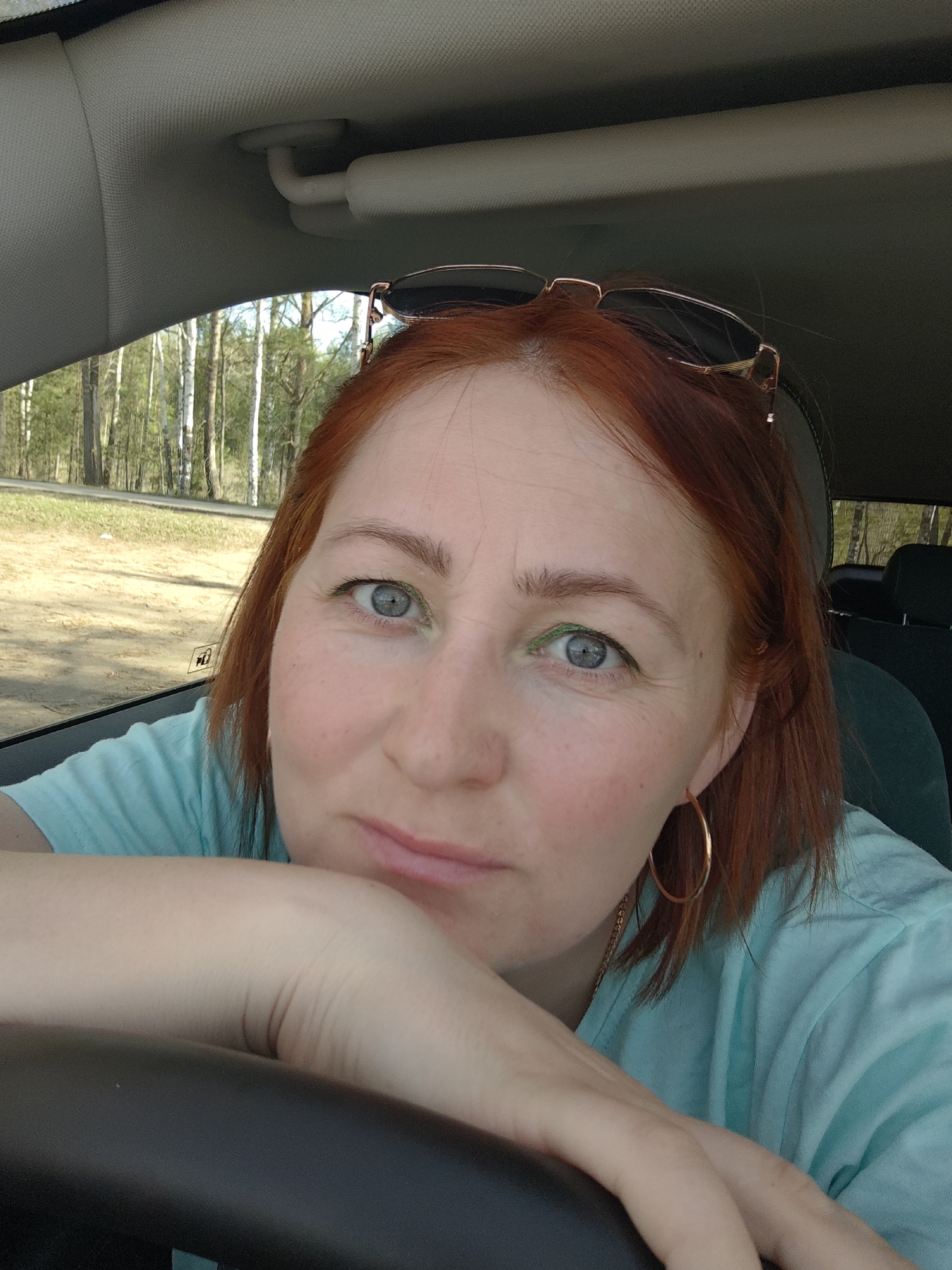 A woman with red hair and blue eyes sits in a car. She wears a light blue t-shirt and gold hoop earrings. Sunglasses rest on top of her head. She leans her arm on the steering wheel. The background shows a forest through the car window. The location is Noviy, Russia.