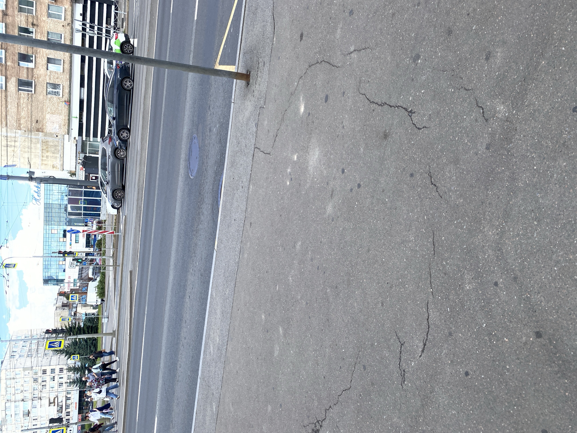 A vertical, eye-level, medium shot captures a street scene in Saint Petersburg, Russia. The foreground is dominated by a grey asphalt sidewalk, showing signs of wear with several cracks and dark spots. A metal pole, likely for signage or a traffic light, stands to the right, emerging from a concrete base on the sidewalk.
The midground features a multi-lane asphalt road. On the opposite side of the road, pedestrians are crossing at a marked crosswalk. They are varied in age and attire, with some dressed casually and others in business-like clothing. A white van is visible behind them, and several cars are parked or in traffic.
The background reveals the urban landscape of Saint Petersburg, with buildings lining the street. These include modern structures with glass facades and older brick buildings. Traffic lights, pedestrian crossing signs with the iconic blue and white pedestrian symbol, and streetlights are visible. The sky is partly cloudy with patches of blue. The overall impression is a typical city street during daytime.