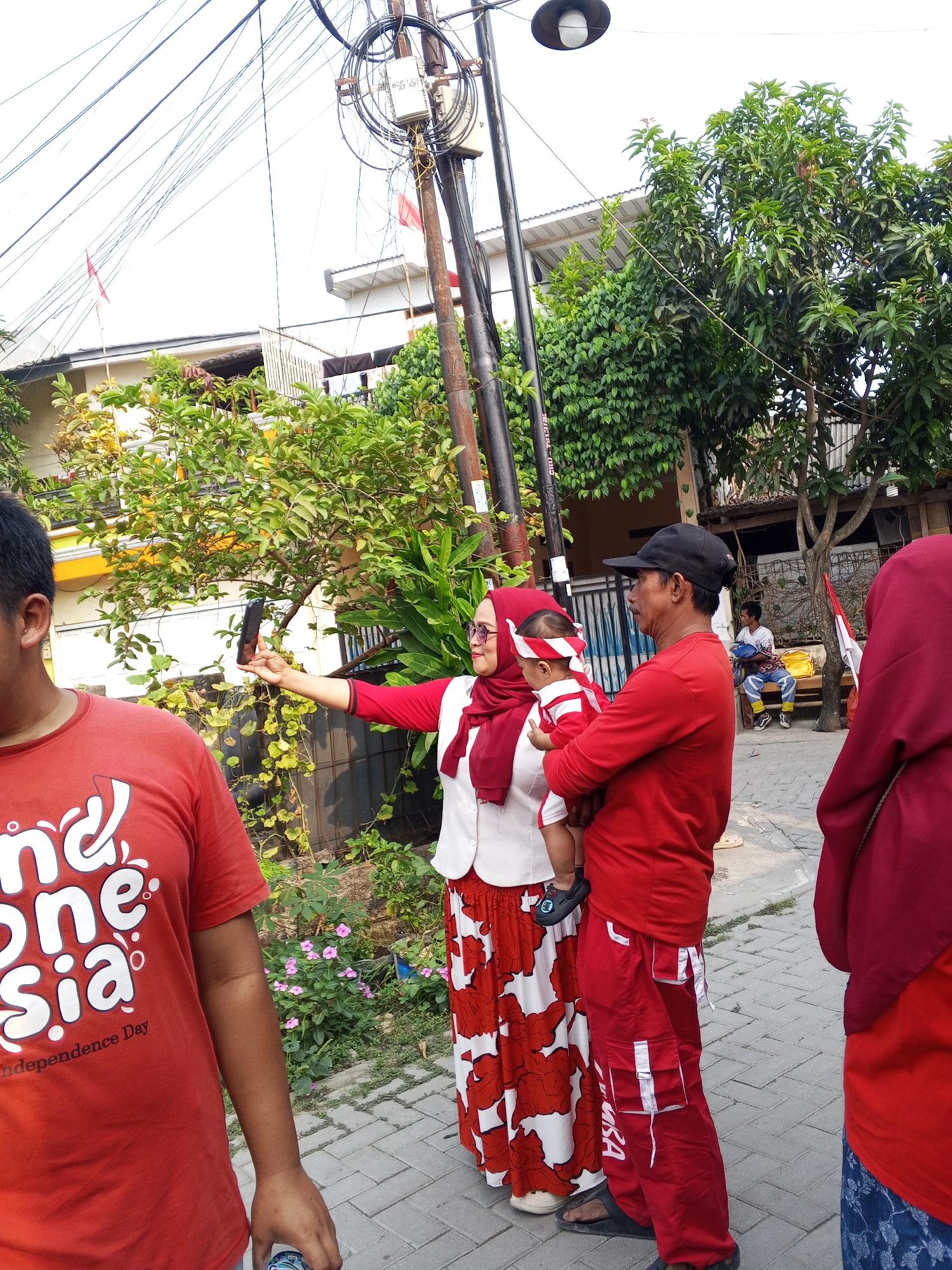 In the city of Ciruas, Indonesia, a street scene unfolds with four individuals dressed in red and white attire, evoking a sense of national pride. A woman, wearing a red hijab, a white vest, and a red and white patterned skirt, stands holding a smartphone in her outstretched left arm, seemingly taking a selfie. She is looking towards the phone, her face partially visible. To her right, a man in a red shirt and red cargo pants, wearing a black baseball cap, holds a baby dressed in red and white striped clothing. The baby has a red and white headband. In the foreground to the left, a person wearing a red t-shirt with "Indonesia" printed on it is partially visible. To the far right, another person in a red hijab is mostly out of frame. The background shows a street lined with buildings, lush green trees, and utility poles with a dense network of wires. A small red and white flag is visible on a pole in the distance, further reinforcing the national theme. The ground is paved with interlocking bricks. The overall atmosphere suggests a casual gathering, possibly related to a national holiday or event.
