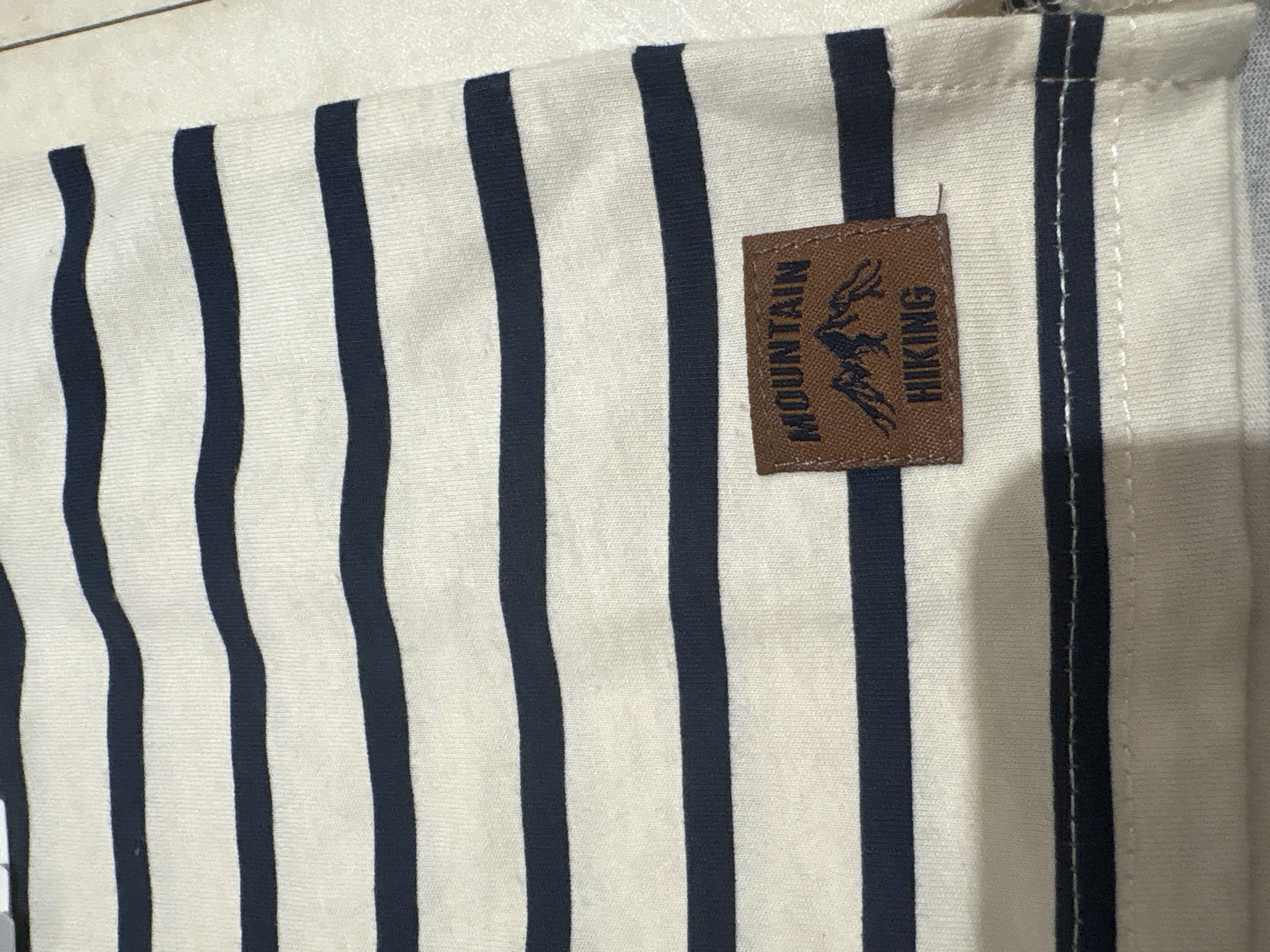 The image shows a close-up of a striped fabric with a brown tag sewn onto it. The fabric has vertical navy blue stripes on an off-white background. The stripes are slightly wavy and vary in thickness. The brown tag is rectangular and has the words "MOUNTAIN HIKING" printed vertically along with an image of what appears to be an eagle. The fabric is partially sewn along one edge, indicated by a line of white stitches.
There is no information in the picture to suggest it was taken in the city of Chaikovsky, Russia.