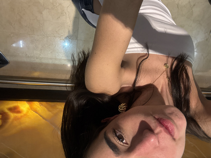 A young adult female is depicted in an indoor setting, posing for a self-portrait. She has dark brown hair, brown eyes, and light skin, with direct eye contact towards the camera. Her head is slightly tilted. She is wearing a white strapless top and dark blue jeans, partially visible. A gold-colored chain necklace with a circular pendant hangs on her chest, and a gold-colored, knot-shaped earring is visible on her right ear. A strong light source illuminates her face and casts shadows on her left arm and chest. The background features a large, rectangular, horizontally oriented panel with an illuminated, amber and orange abstract pattern, likely a backlit stone slab, framed in a dark material. Below this panel, a horizontal metallic railing is visible. The floor is composed of polished light-colored marble or tiles, exhibiting reflections. A metallic or silver object is partially visible in the lower right corner of the frame. The location is an interior space within the city of Unknown, Unknown, likely a public or semi-public establishment.