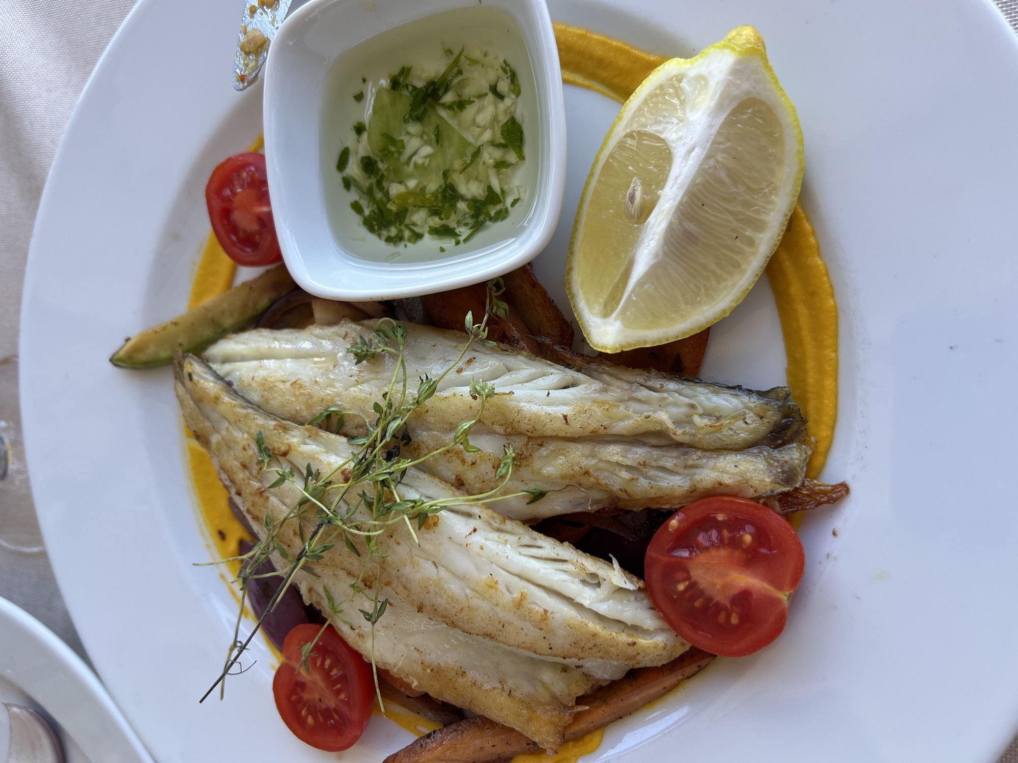 A white plate of grilled fish is presented in a bright, natural light setting, likely an outdoor dining area given the blurred background which suggests a patio or terrace. The dish features two fillets of white fish, lightly browned and seasoned, placed atop a vibrant yellow puree, possibly pumpkin or carrot. Accompanying the fish are several small, halved cherry tomatoes, a wedge of fresh lemon, and what appears to be a small dish of dressing containing finely chopped herbs and garlic. Some roasted vegetables, possibly carrots and zucchini, are also visible underneath the fish. A sprig of fresh thyme garnishes the fish. The plate rests on a light-colored tablecloth. No people are visible in the frame. The overall impression is of a fresh, well-prepared meal.