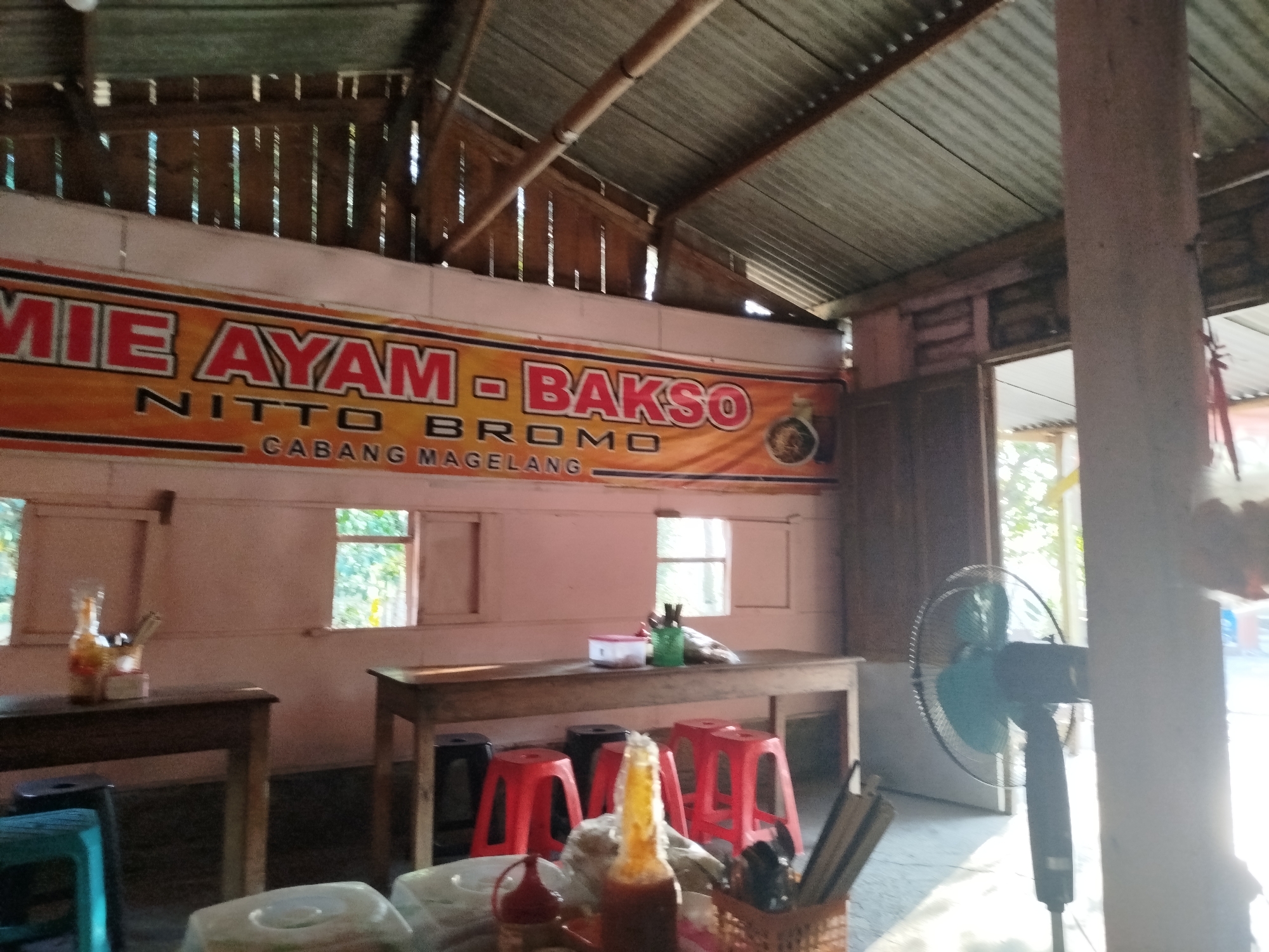 The image shows the interior of a simple restaurant, likely a "warung" or roadside eatery, in Pojok, Indonesia. A large banner above the main dining area prominently displays "MIE AYAM - BAKSO NITTO BROMO CABANG MAGELANG," indicating the establishment's name and specialization in chicken noodles and meatballs, with a branch in Magelang.
The restaurant appears rustic, with a corrugated metal roof supported by wooden beams and vertical wooden slats. The walls are painted a pale pink. Several wooden tables are set up for dining, some with red and black stools scattered around them. One table on the left has a bottle with a yellow liquid and a holder containing utensils. Another table in the center has a container with a red lid and a green holder with more utensils and what appear to be plastic bags.
In the foreground, a bottle of sauce is visible, along with a basket containing cutlery. To the right, a standing electric fan is positioned near an open doorway that leads to an outdoor area, with bright sunlight filtering in. The overall impression is of a functional, no-frills eating establishment catering to local patrons. There are no people visible in the frame.