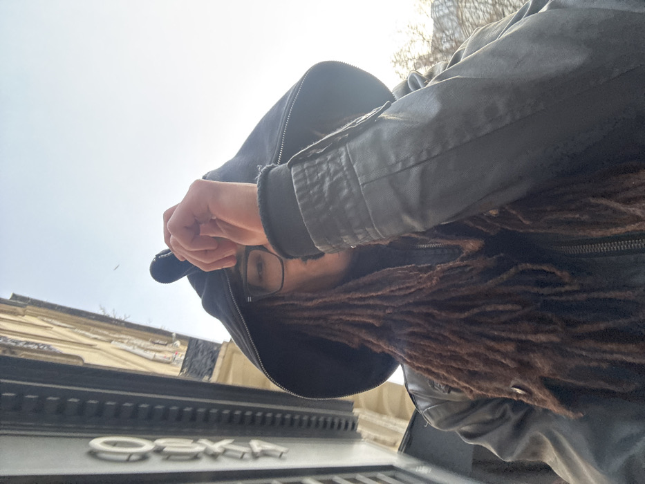 A person in a hooded jacket is angled downward, obscuring much of their face. Only their eyes and eyeglasses are visible, along with dreadlocks that cascade forward. Their hand is raised near their face. The sky is pale and visible in the background. In the foreground, below the person, a building with raised lettering, reading "OSKA," is partially visible. The image is tilted, creating a disorienting perspective.