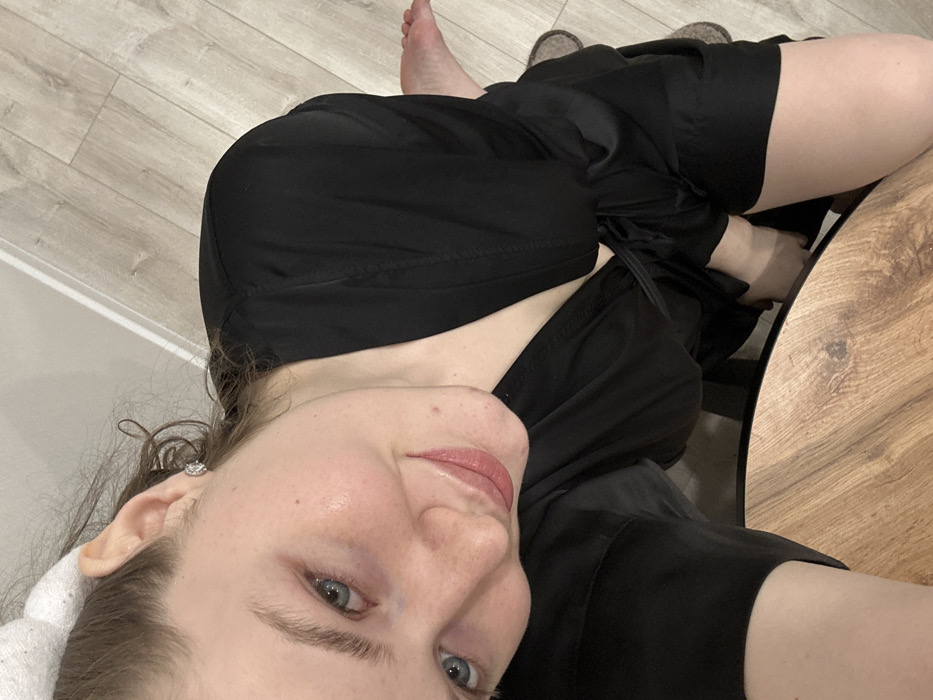 A woman is pictured in a selfie, her face turned towards the camera with a slight smile. She wears a black silky robe, tied at the waist. A white headband sits on her head, holding back her hair. One earring is visible. Her legs, clad in the same black material, rest on a wooden table, while her bare feet are visible near the floor. The background includes a section of light-colored wood flooring.