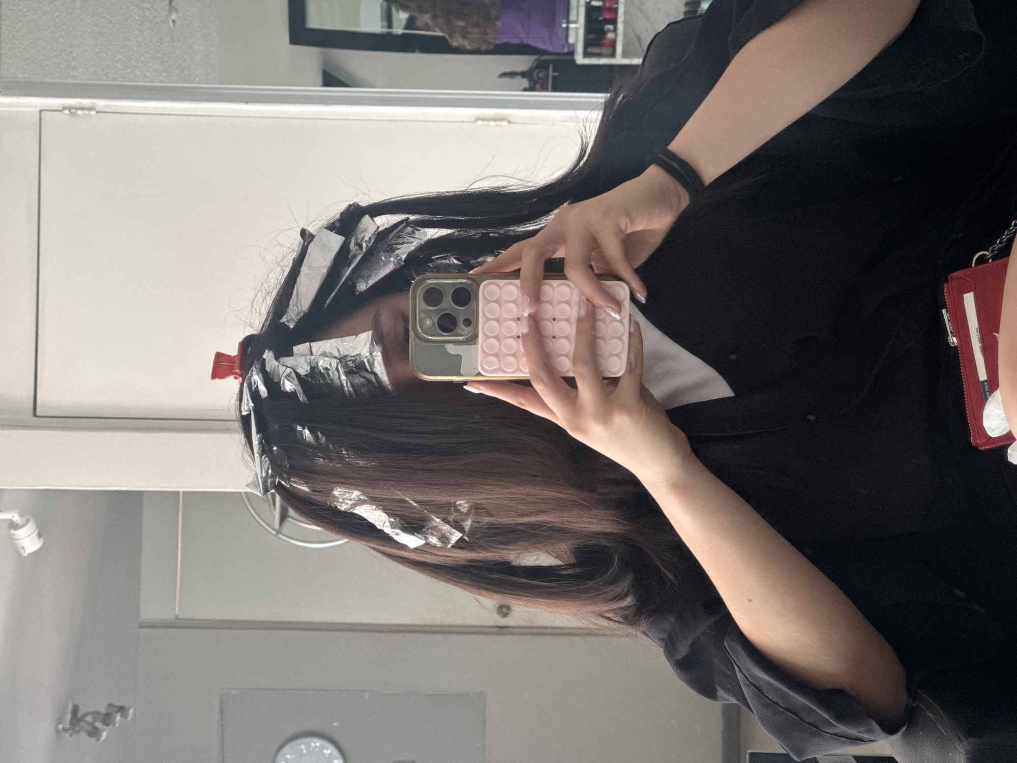 A person is seated in what appears to be a hair salon in Puebla, Mexico, based on the available information. The person is having their hair highlighted, as evidenced by the aluminum foil and red clip in their hair. They are holding a smartphone in front of their face, likely taking a selfie in the mirror. The phone has a distinctive pink "pop-it" style case. The person is wearing a black garment. In the background, part of the salon environment is visible, including a mirror, a white door or wall, and possibly hair products on shelves. A red wallet or small purse is also visible.