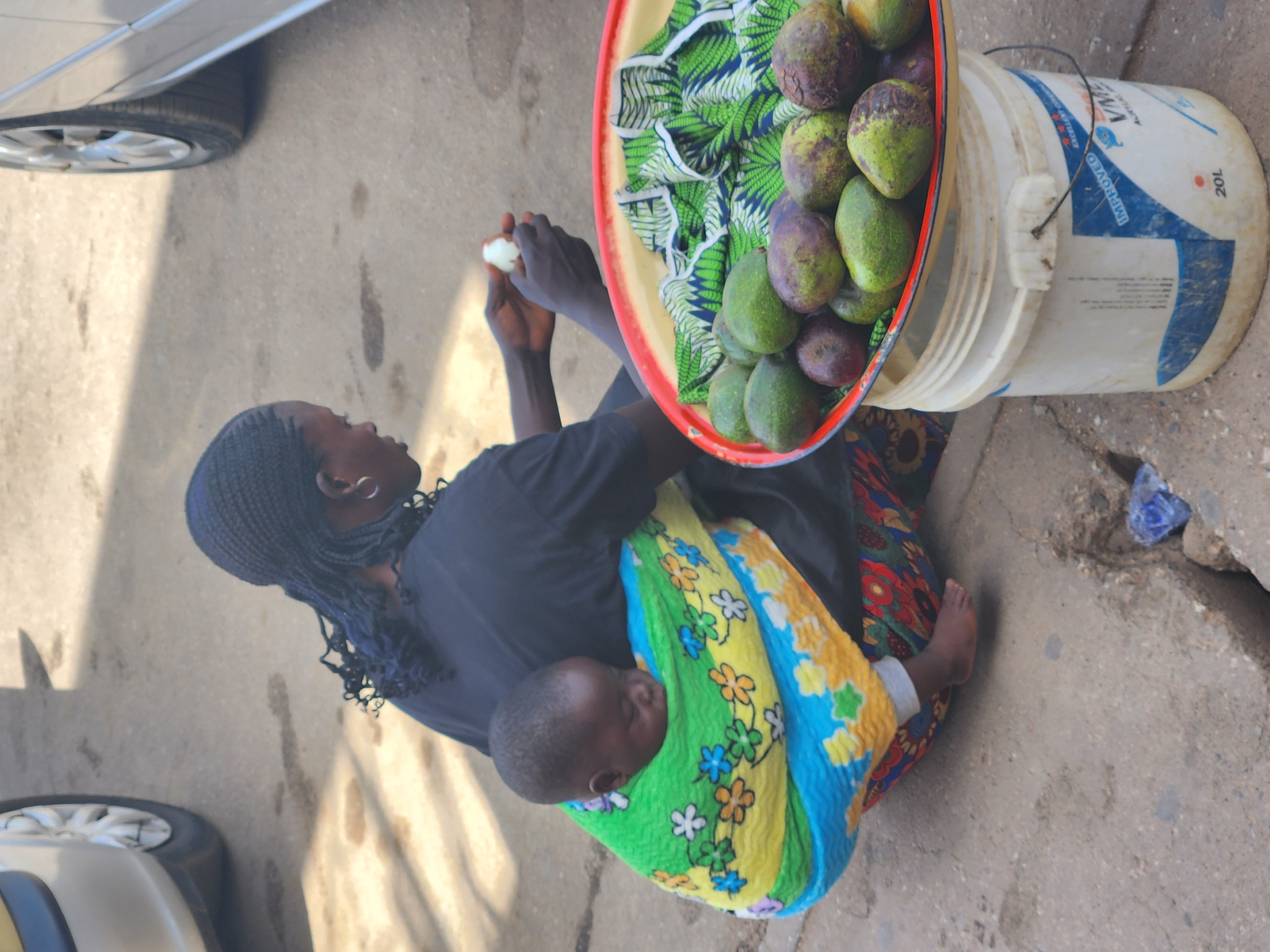 This photograph captures a woman and a baby in an outdoor setting, likely a roadside or market in Jos, Nigeria. The woman is seated on concrete ground, wearing a dark top and a vivid, multi-colored patterned skirt. Her hair is styled in dark braids, and she wears hoop earrings. She is actively engaged in handling a small, light-colored item with both hands, possibly peeling or preparing it.
A baby is visible on her back, securely wrapped in a colorful blanket featuring green, yellow, and blue sections adorned with floral patterns. The baby appears to be asleep.
Adjacent to the woman is a large, red-rimmed tray filled with numerous avocados, which exhibit a range of green and purplish-brown hues. A green and white patterned cloth is also present in the tray. The tray is supported by a white plastic bucket. The bucket clearly displays "20L" and some other partially visible text, including "IMPROVED".
In the background, on the left side, the tires and lower sections of two vehicles are partially visible, suggesting a location near a road or parking area. The strong shadows on the ground indicate it is daytime, likely a sunny day. The scene depicts a common sight of street vending, with the woman attending to her goods while carrying her child.