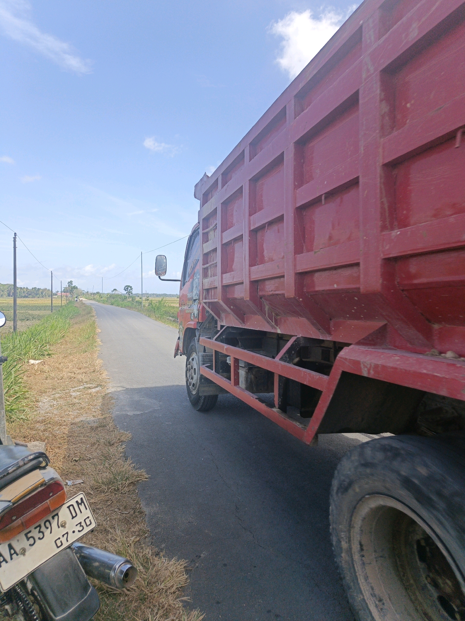 The image depicts a rural roadside scene in Kamulyan, Indonesia. A large, red dump truck is parked on the right side of an asphalt road, partially obscuring the view of a motorcycle in the foreground on the left. The truck is oriented towards the right of the frame, with its side facing the camera. Its cargo bed is a prominent feature, showing a structural design with horizontal and vertical beams. The motorcycle's license plate, displaying "AA 5397 DM" and "07-3G", is visible in the lower left corner. The road continues into the distance, bordered by dry, grassy fields on both sides. Sparse vegetation, including some taller grass and scattered trees, is visible further down the road and in the background, against a bright blue sky with scattered white clouds. Utility poles with overhead wires are present along the roadside. The overall impression is that of a sunny day in a quiet, agricultural area.