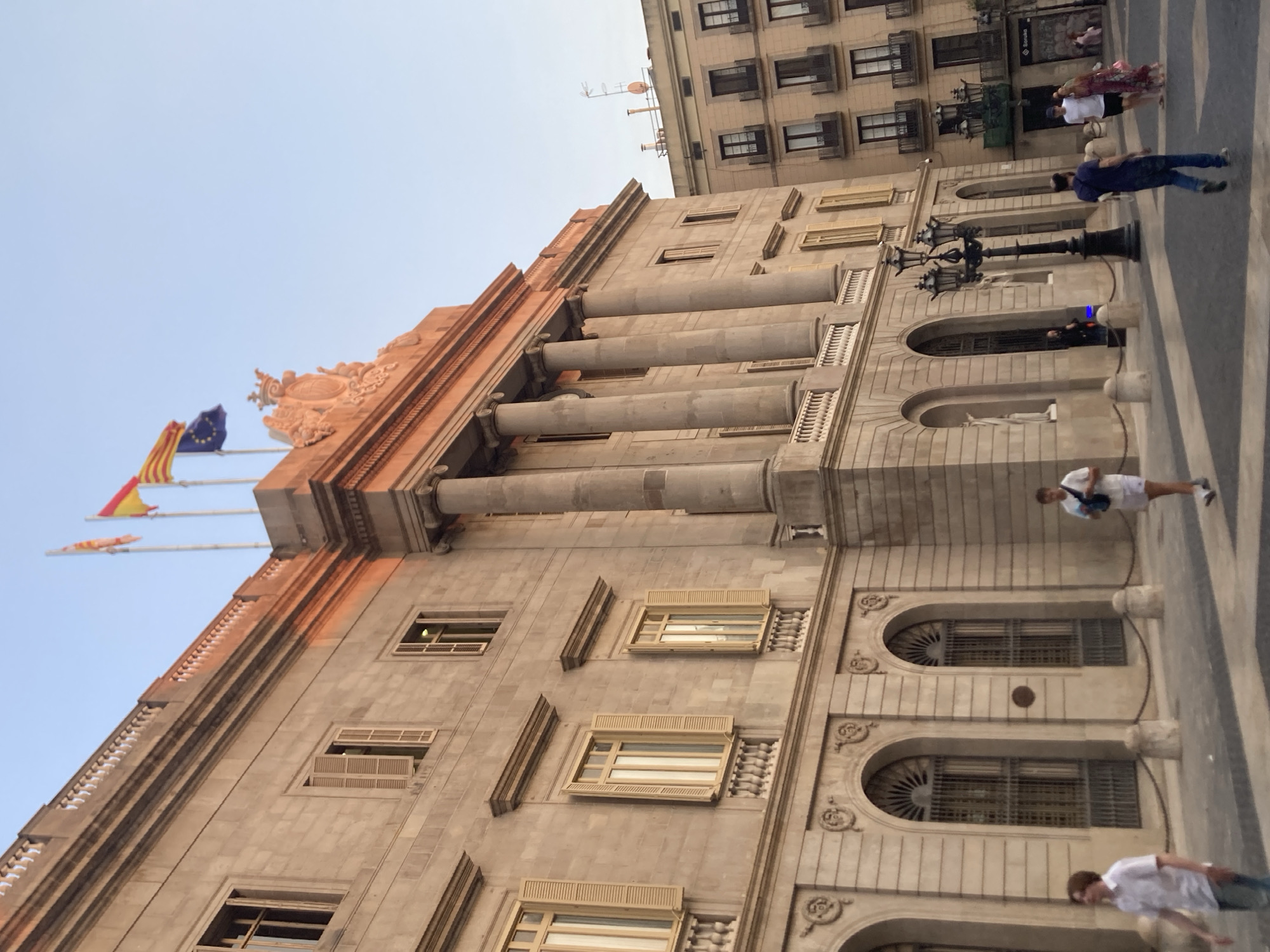 The image depicts the Palau de la Generalitat de Catalunya, a historical building located in Barcelona, Spain. The facade of the building is made of stone and features classical architectural elements such as columns and arched windows. Three flags are visible atop the building: the Catalan flag, the Spanish flag, and the European Union flag. In the foreground, several people are walking on a paved plaza in front of the building. The sky is clear and blue. The overall scene is tranquil, with a mix of historical architecture and everyday life.