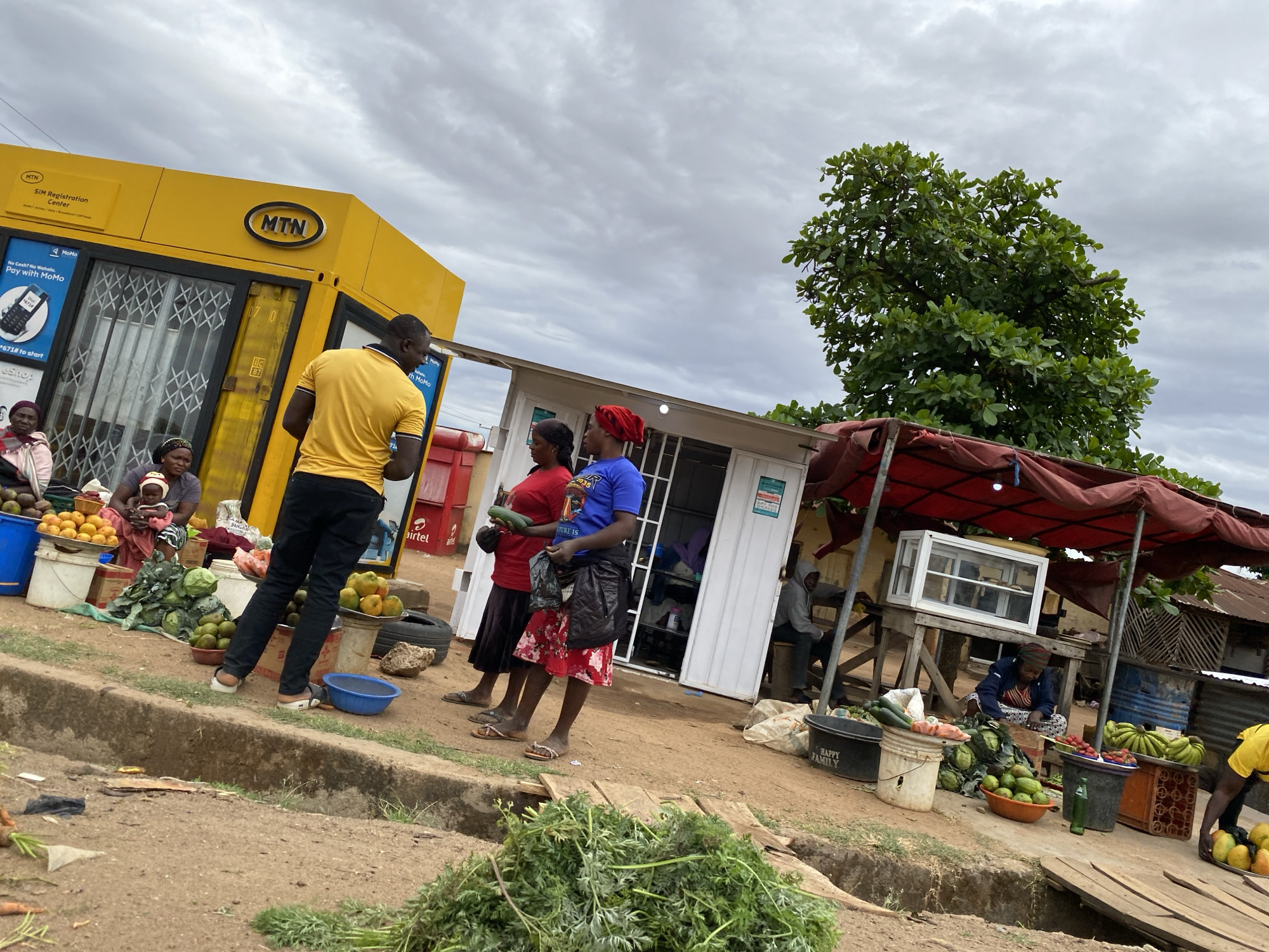 The image shows a bustling outdoor market scene in Bukuru, Nigeria, under a cloudy sky. In the foreground, a pile of green leafy vegetables sits beside a dirt path separated by a concrete curb. A man in a yellow shirt and black pants stands near a yellow MTN kiosk where two women are seated. One woman holds a baby. Further back, two women stand near a white kiosk, one holding a cucumber. A red Airtel sign is visible between the MTN and white kiosks. To the right, a market stall with a red awning displays produce. A woman in a blue and white patterned headwrap is seated among the produce. Another person in a gray hoodie sits behind the stall. Buckets, crates, and other containers hold a variety of fruits and vegetables, including mangoes, avocados, cabbages, tomatoes, and bananas.