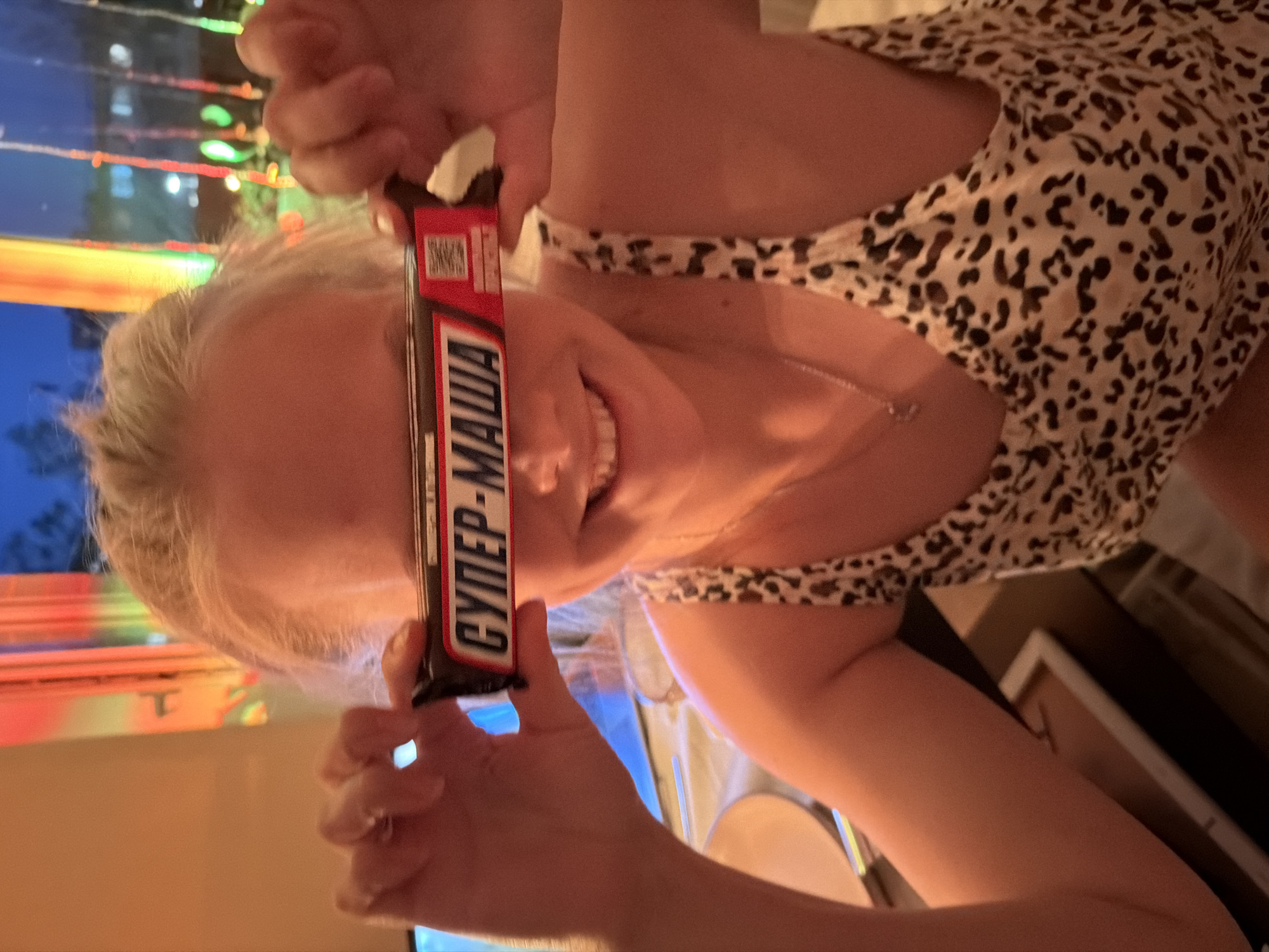 A person with blonde hair is holding a "Супер-Маша" candy bar in front of their face. The person is wearing a leopard-print top. The background appears to be a room with a window showing a cityscape at night. The image may have been taken indoors in Izhevsk, Russia based on the product name on the candy bar.