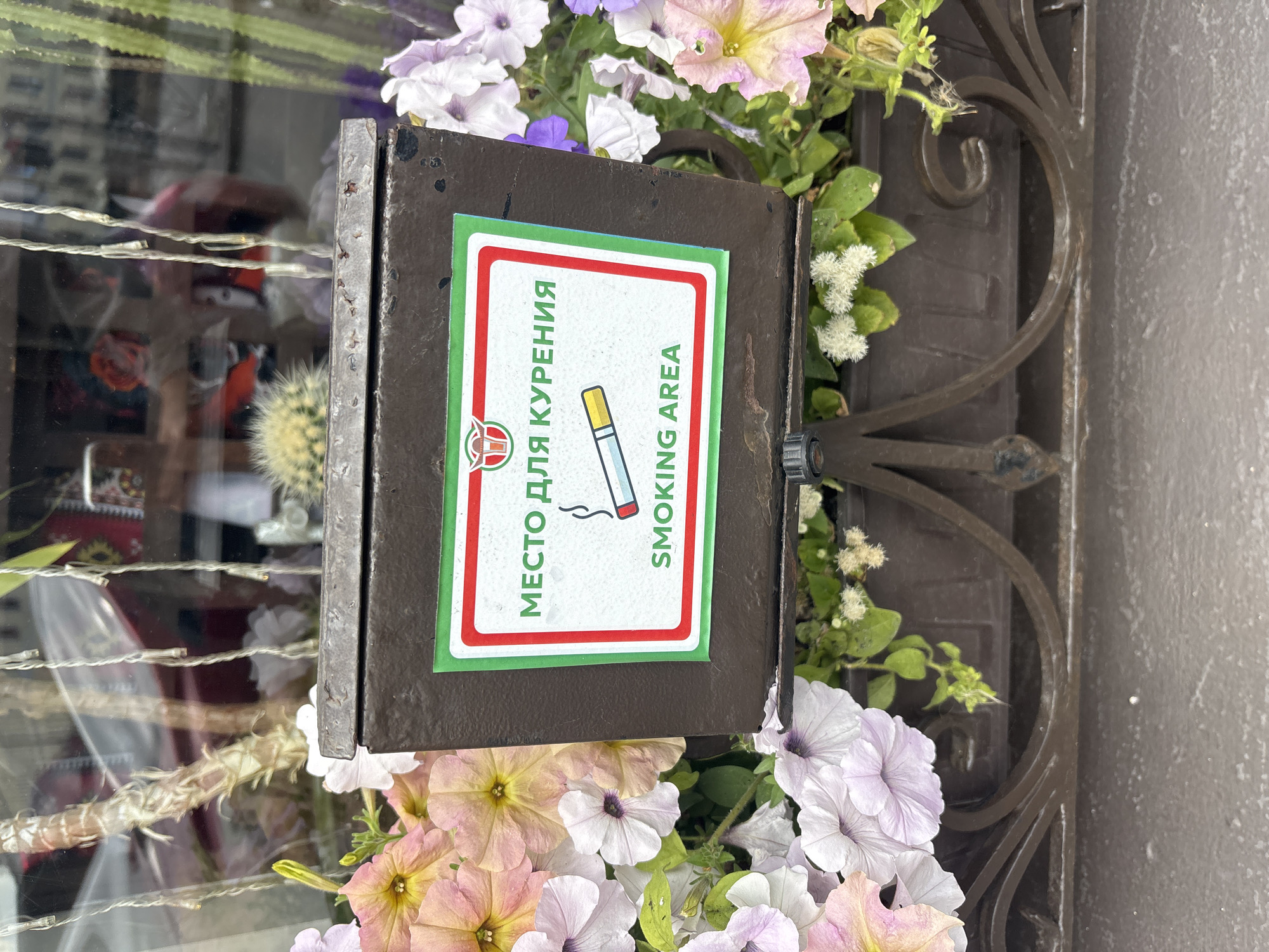 The image shows a dark brown metal ashtray container, possibly made of cast iron, with a sign attached. The sign has a white background with a red and green border. At the top of the sign is a red logo featuring a stylized bull's head. Below the logo, in green Cyrillic text, it reads "МЕСТО ДЛЯ КУРЕНИЯ" which translates to "SMOKING AREA". Below this, in green English text, it also reads "SMOKING AREA". A graphic of a lit cigarette is centered on the sign below the text. The ashtray is surrounded by a planter box filled with pink, white, and purple petunias. In the background, reflections in a window show glimpses of buildings, lights, and possibly people. The overall scene appears to be outdoors, likely on a city street. The presence of Cyrillic text suggests a location in a Russian-speaking country, and the style of the architecture visible in the reflection could indicate Saint Petersburg, Russia.
